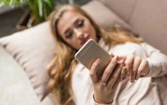 A woman with long hair is lying on a couch, holding and looking at a smartphone. Her focus is on the screen, and she is resting on a cream-colored pillow. A plant is visible in the background.