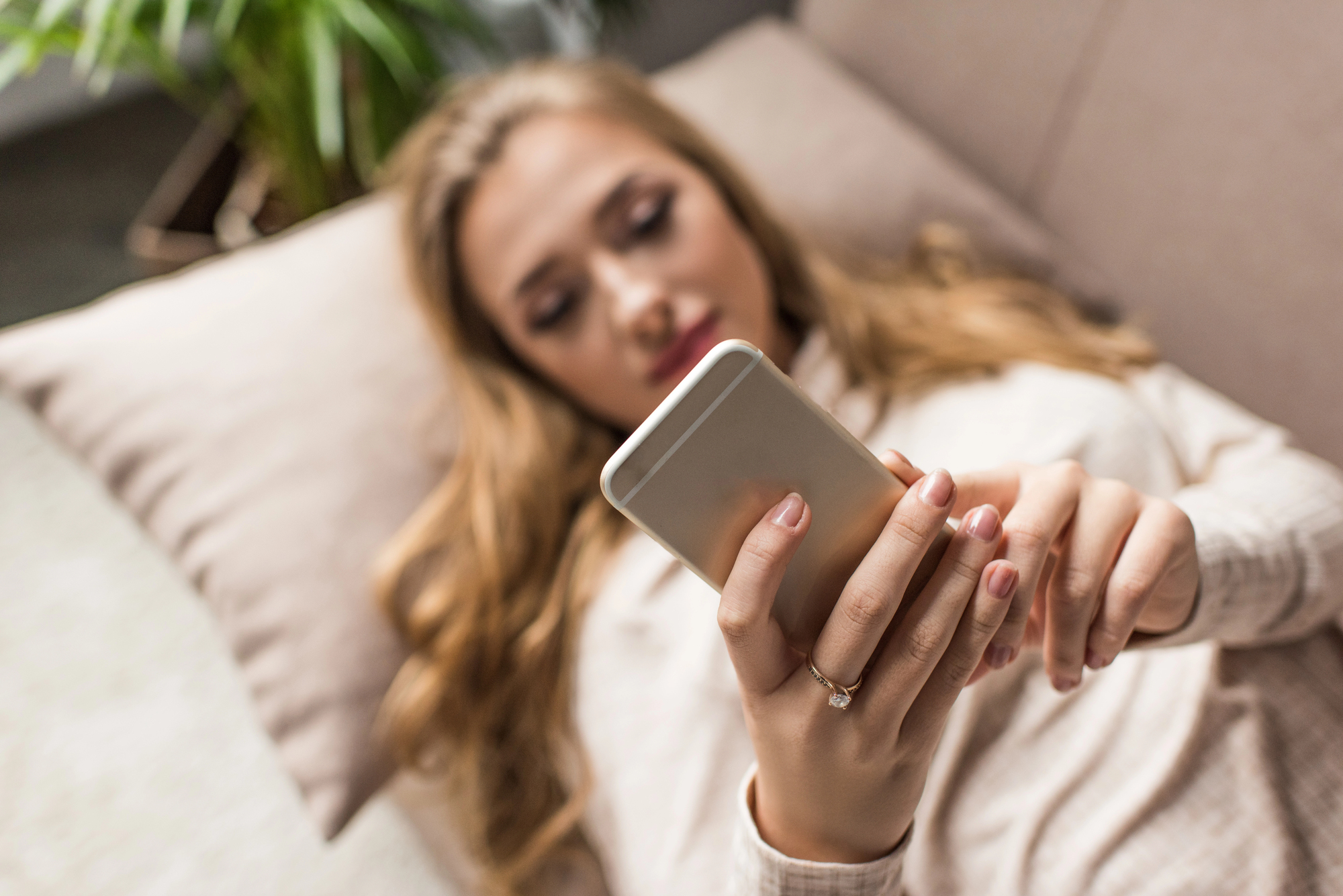 A woman with long hair is lying on a couch, holding and looking at a smartphone. Her focus is on the screen, and she is resting on a cream-colored pillow. A plant is visible in the background.