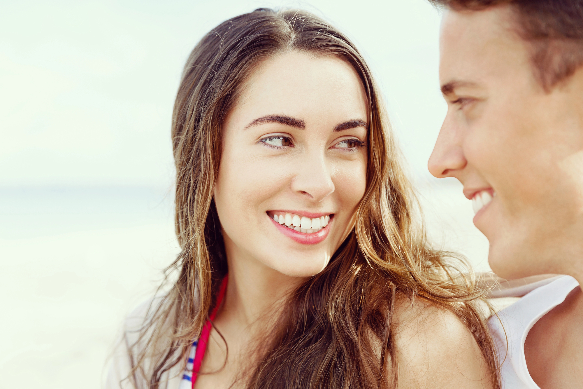 A close-up of a woman and a man smiling at each other while sitting on a beach. The woman has long brown hair and is wearing a bright swimsuit, while the man is wearing a light shirt. They appear to be enjoying a sunny day.