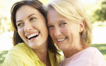 A close-up of two women smiling outdoors. One has long brown hair and wears a yellow top, while the other has short blonde hair and wears a pink top. They are embracing and appear cheerful, with greenery in the background.