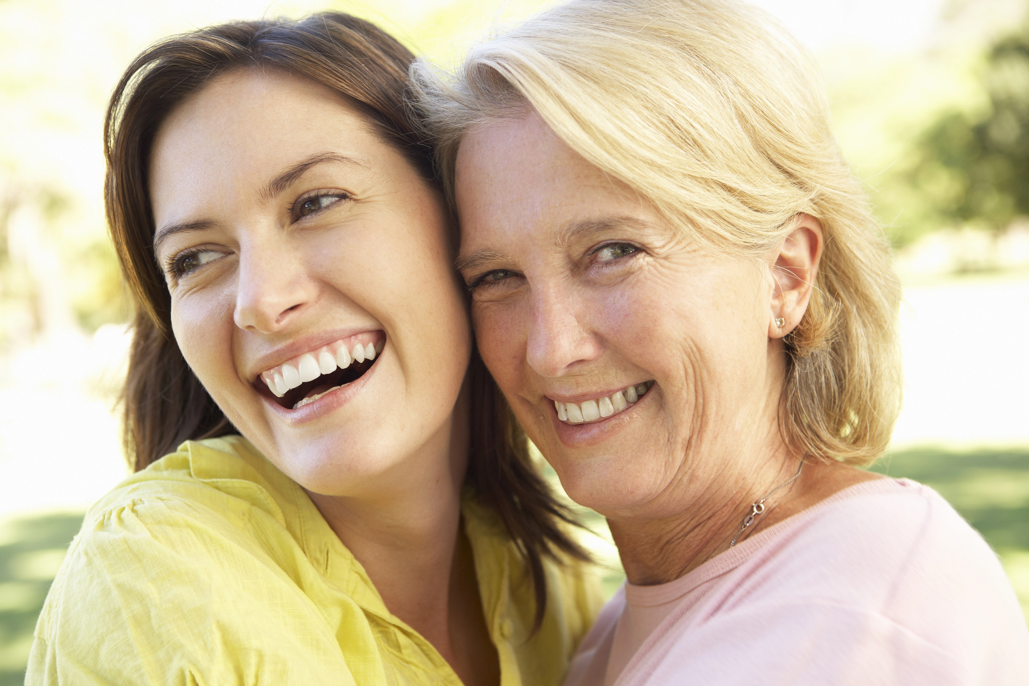 A close-up of two women smiling outdoors. One has long brown hair and wears a yellow top, while the other has short blonde hair and wears a pink top. They are embracing and appear cheerful, with greenery in the background.