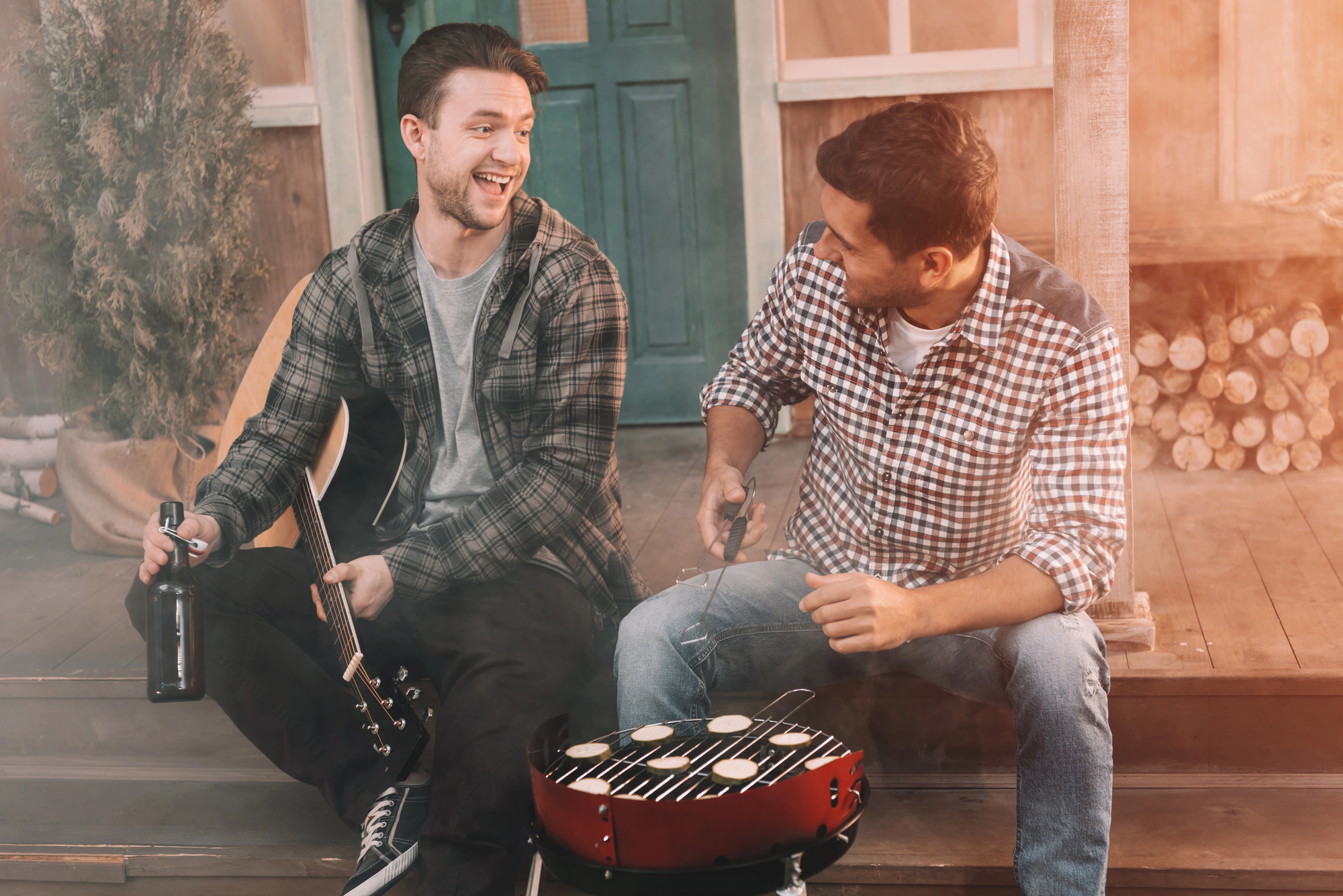 Two men sitting on a porch, smiling and chatting. One holds a beer and a guitar, while the other tends to food on a small grill. The setting is relaxed, with logs and greenery in the background. Sunlight adds a warm ambiance.