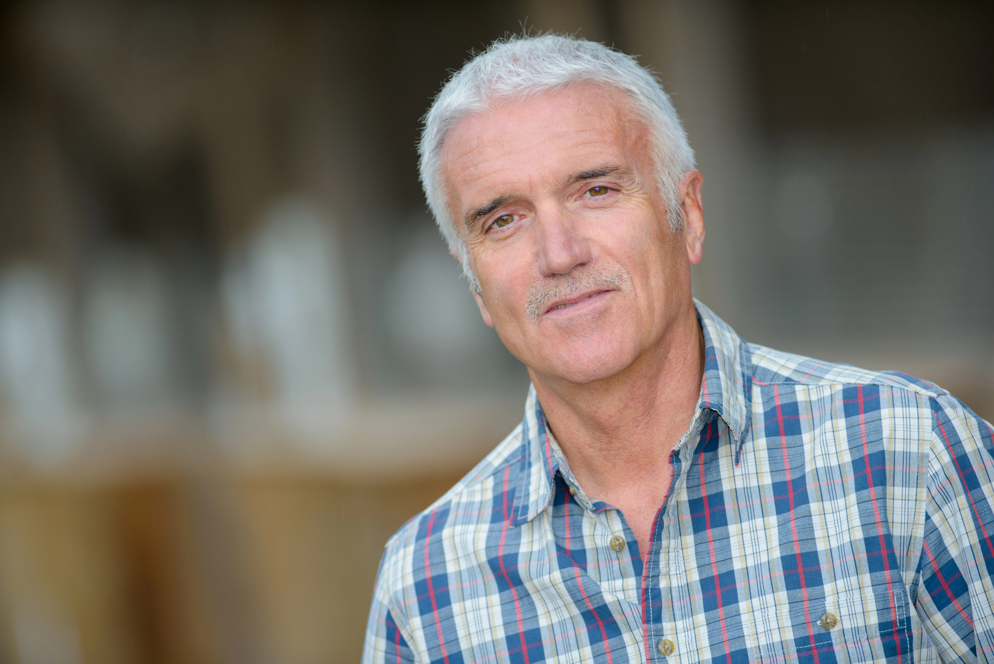 Older man with short white hair and a mustache, wearing a plaid shirt, smiling softly at the camera. The background is blurred.