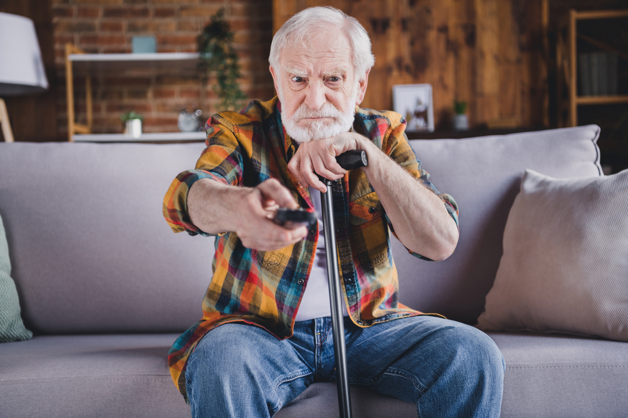 An elderly man with a white beard sits on a gray couch in a cozy living room, holding a remote control in one hand and a walking cane in the other. He wears a colorful plaid shirt and jeans, gazing forward intently.