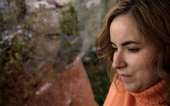 A woman in an orange sweater looks out of a rain-speckled window. Her reflection is visible on the glass, merging with the view of the rainy exterior.