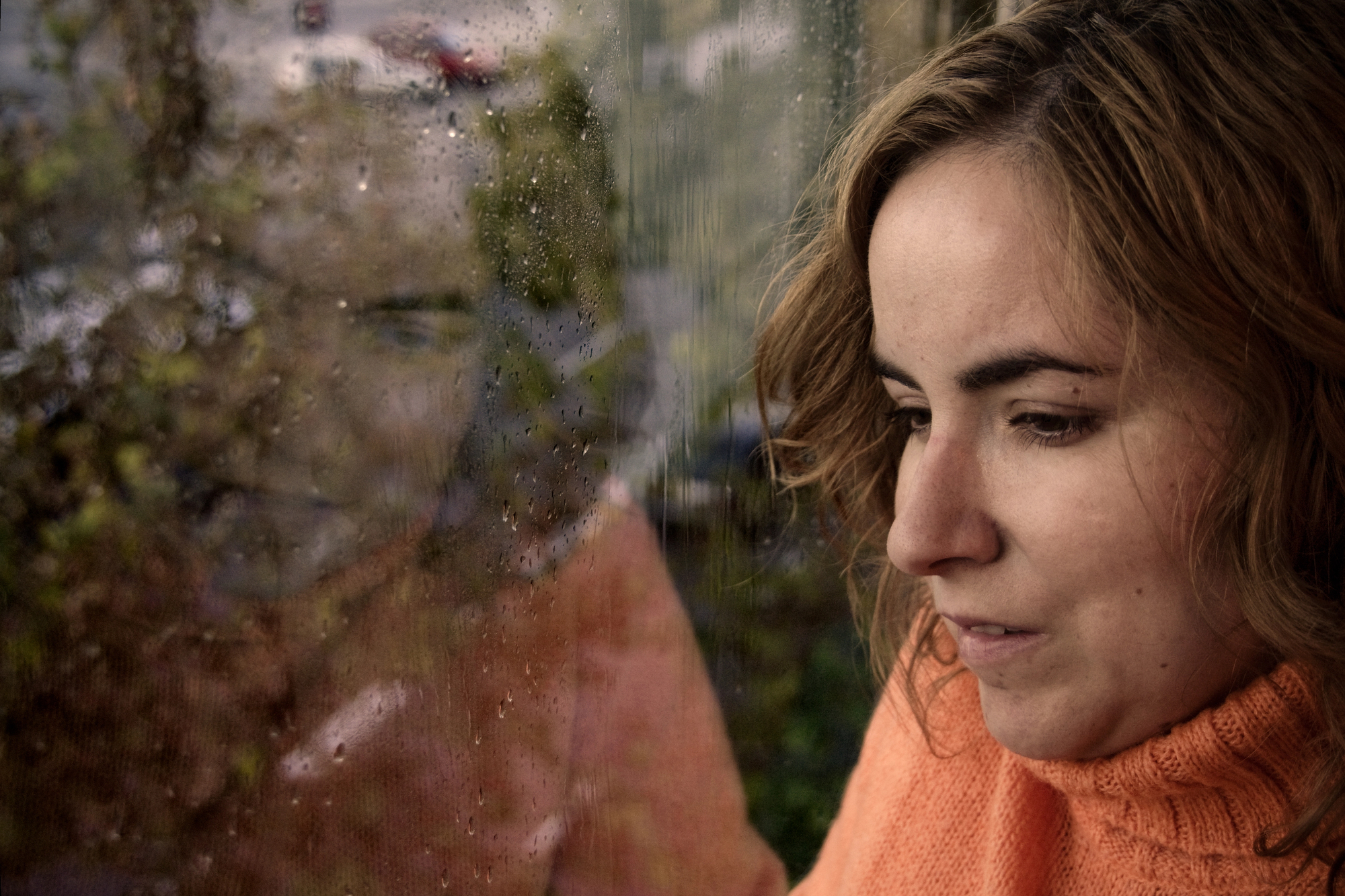 A woman in an orange sweater looks out of a rain-speckled window. Her reflection is visible on the glass, merging with the view of the rainy exterior.