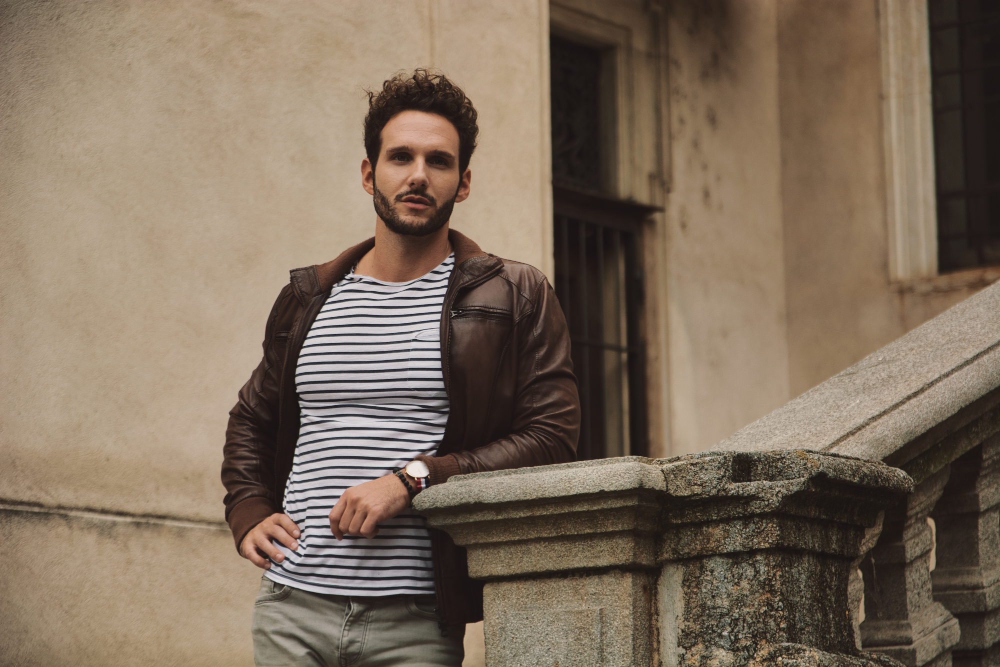 A man with curly hair leans casually against an outdoor stone staircase. He is wearing a brown leather jacket over a striped shirt and olive pants. The background features a weathered beige wall with a window.