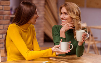 Two women sit at a wooden table, both smiling and holding white mugs. One wears a yellow sweater, the other a green one. A smartphone rests on the table between them. The background features brick walls and warm indoor lighting.