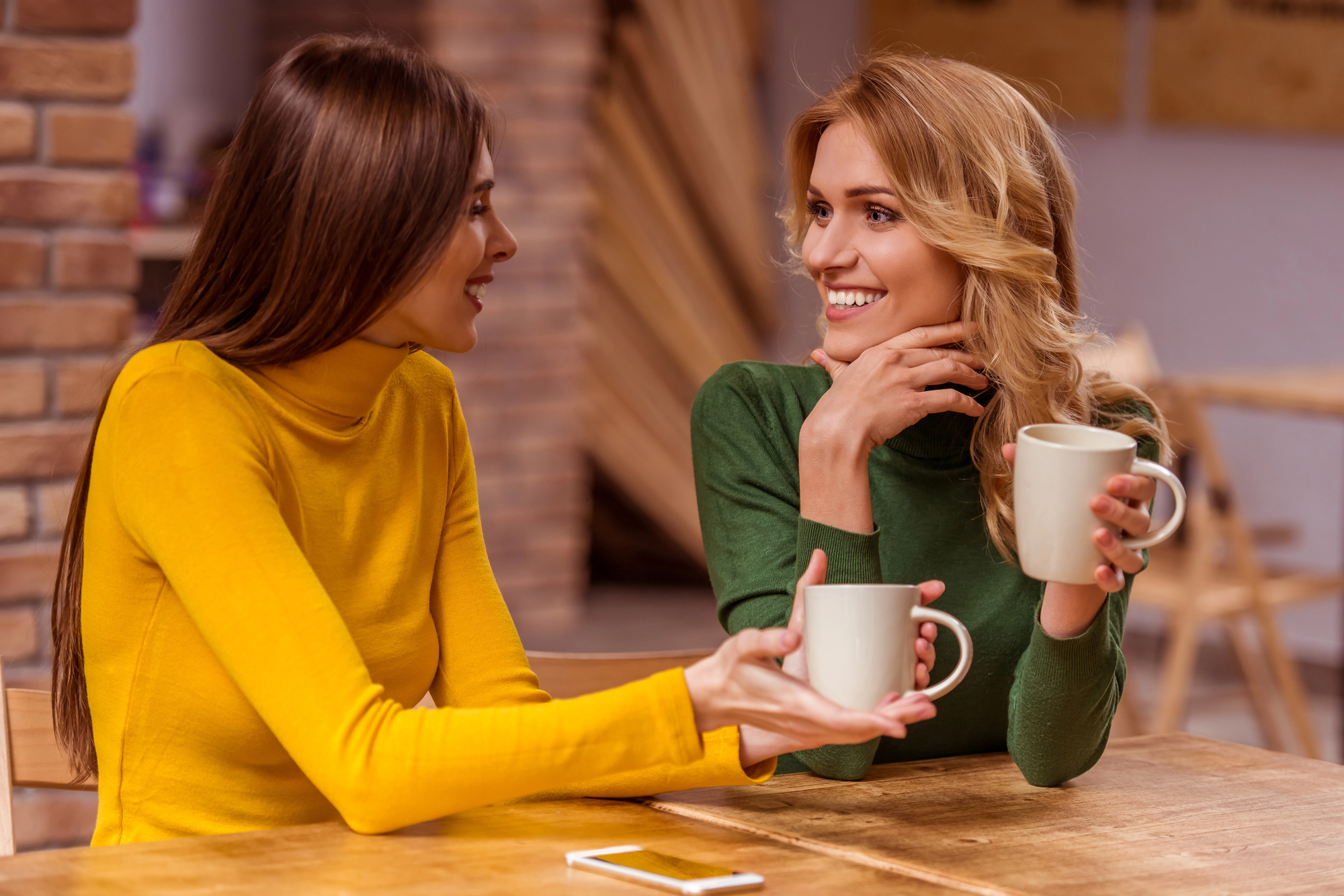 Two women sit at a wooden table, both smiling and holding white mugs. One wears a yellow sweater, the other a green one. A smartphone rests on the table between them. The background features brick walls and warm indoor lighting.