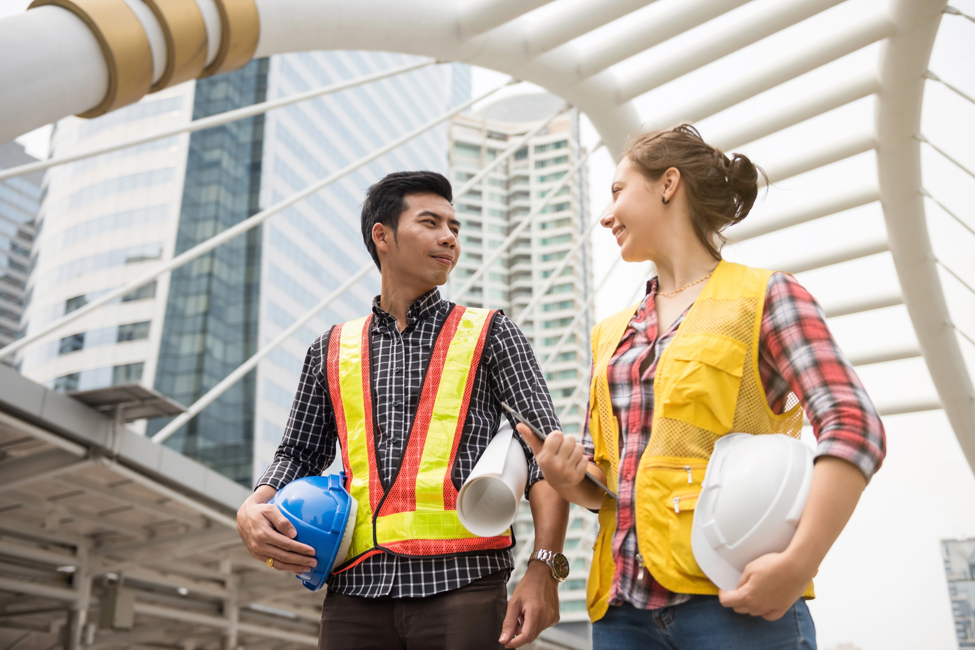 A man and woman in safety vests stand outdoors in an urban setting, holding hard hats and architectural plans. They appear to be discussing a construction project, with modern high-rise buildings in the background.