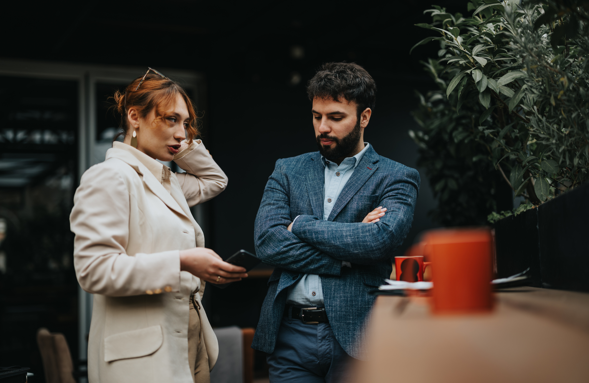 A woman in a beige coat looks at her phone while speaking to a man in a blue blazer with crossed arms. They are outdoors near some plants, with a red mug visible on a table beside them.