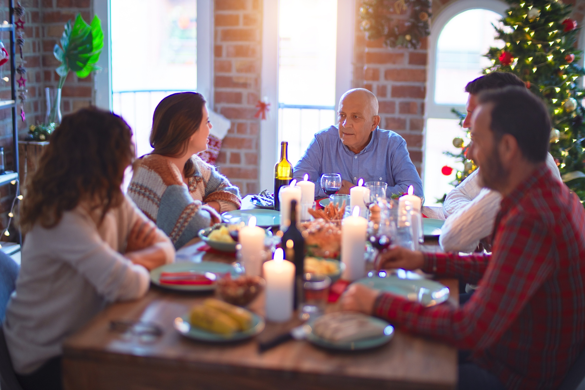 A group of people sits around a wooden dining table, sharing a meal in a warmly lit room with a decorated Christmas tree in the background. Candles are lit on the table, creating a cozy atmosphere.