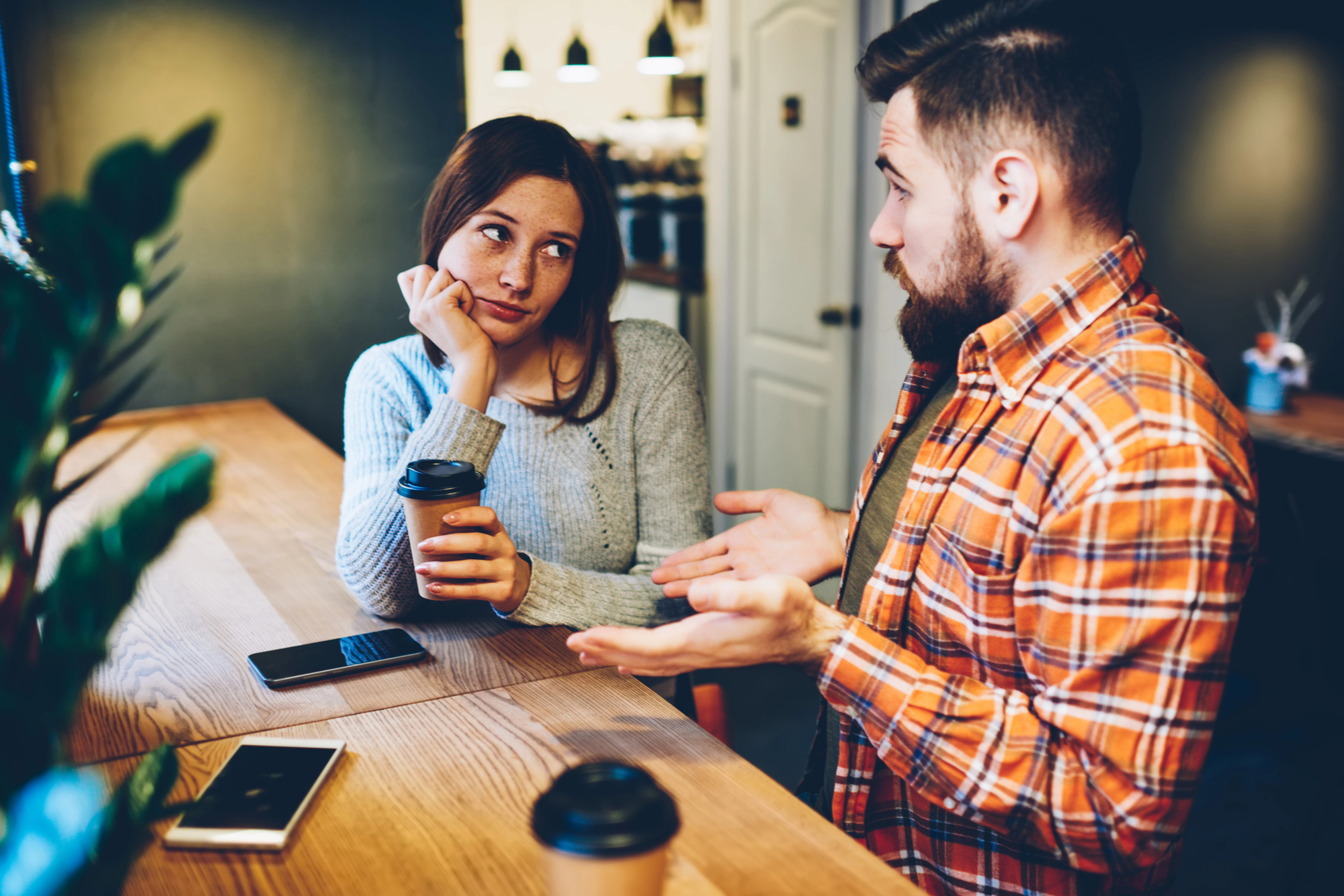 A man and woman sit at a wooden table with coffee cups and phones. The man, wearing a plaid shirt, gestures while talking. The woman, in a gray sweater, listens with a thoughtful expression, her head resting on her hand. The setting appears to be a casual café.