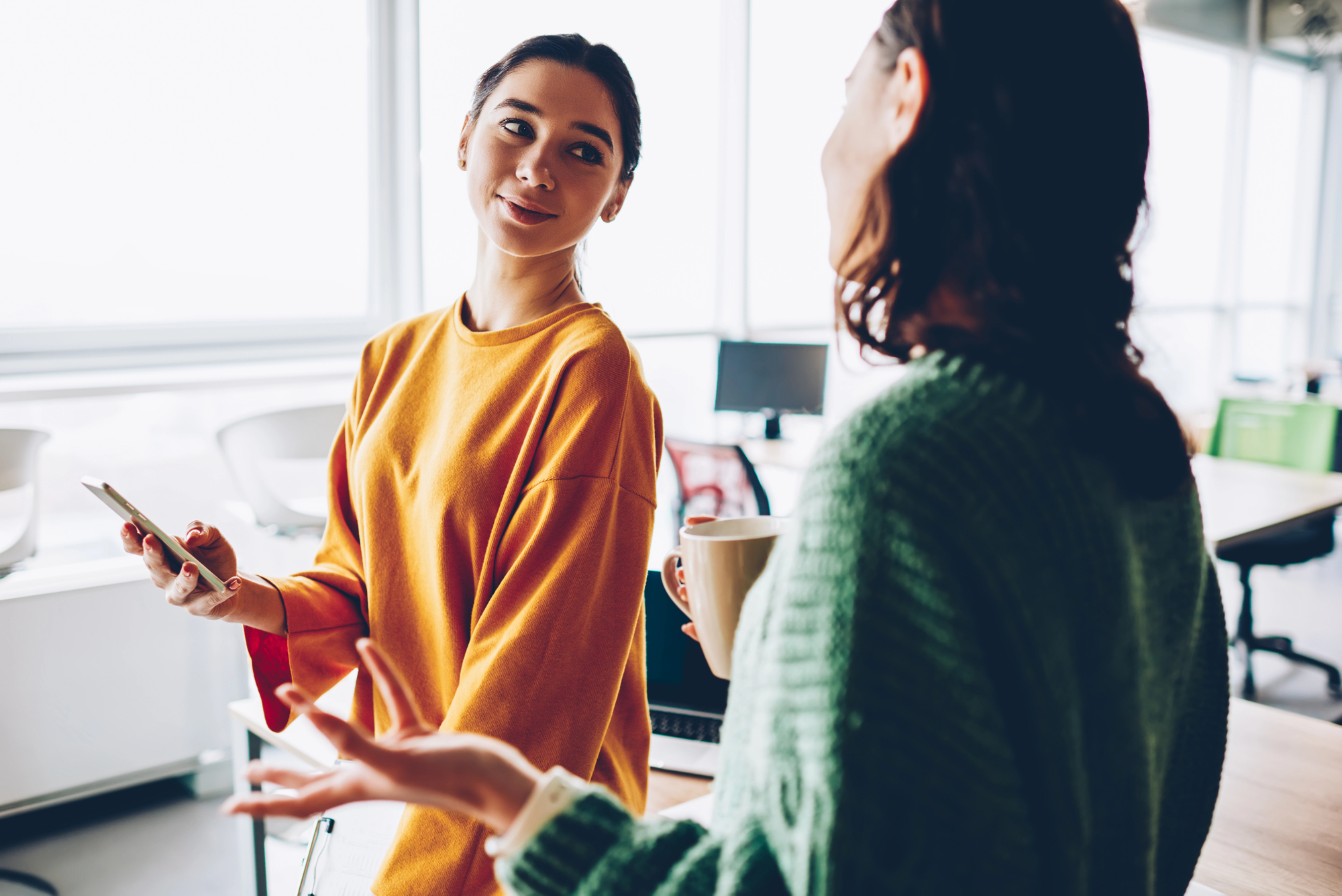 Two women are conversing in a bright office. One is holding a smartphone and wearing an orange sweater, while the other has a mug and a green sweater. They appear engaged in a friendly discussion, with computers in the background.