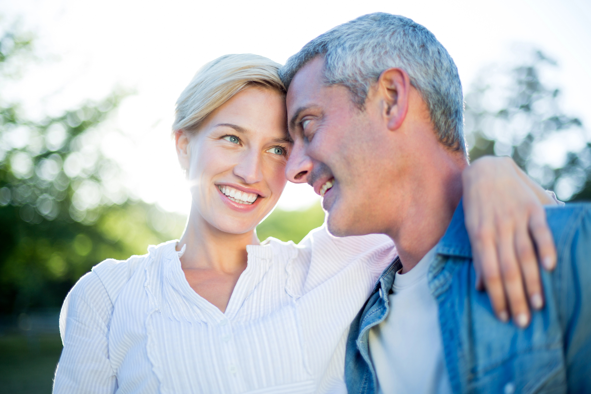 A smiling couple stands close together outdoors. The woman has short blonde hair and wears a white blouse, while the man has short gray hair and wears a denim shirt. They appear happy and relaxed amidst a backdrop of trees and sunlight.