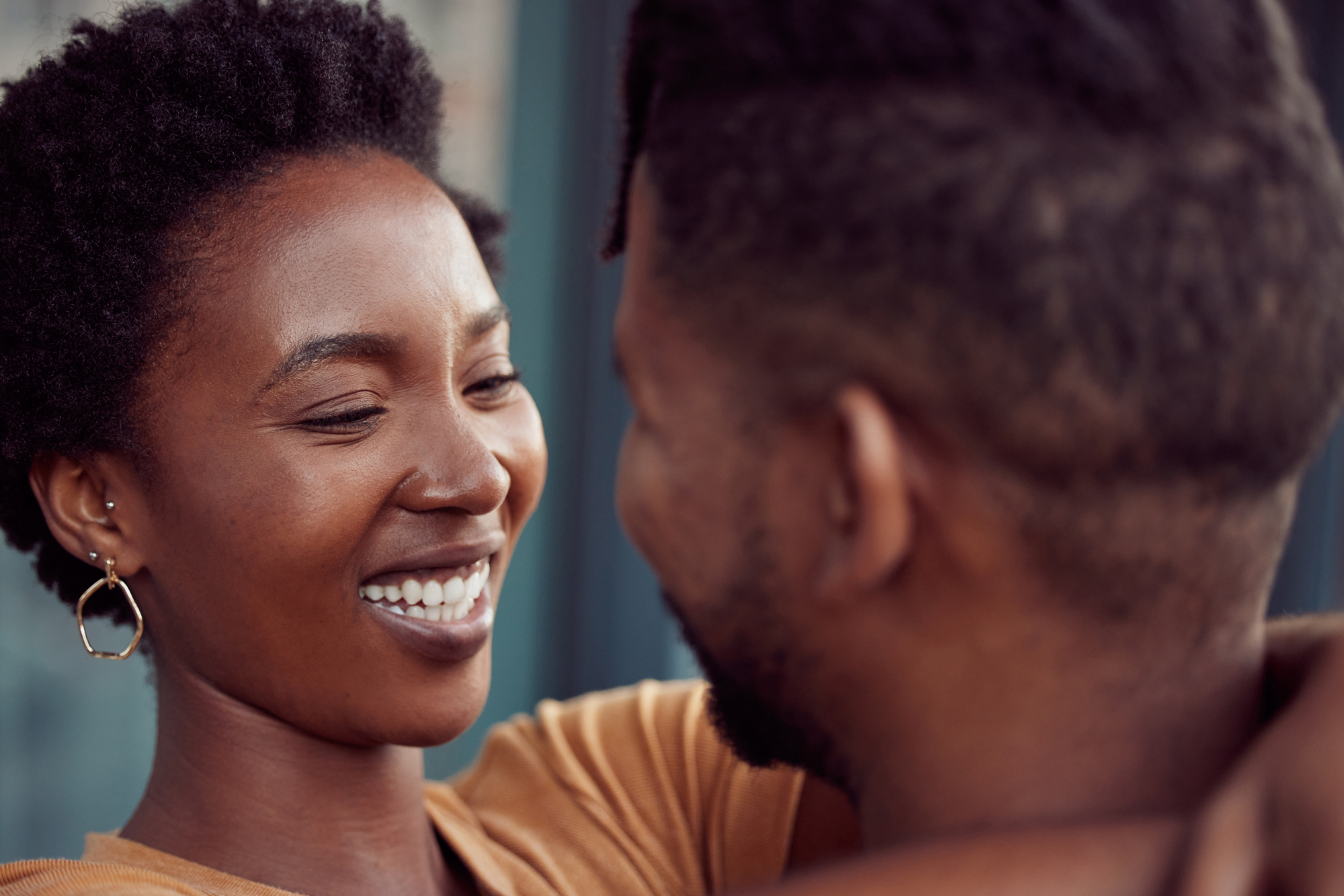 A smiling woman with short curly hair and hoop earrings looks affectionately at a man with short hair. They are embracing closely, set against a blurred background.