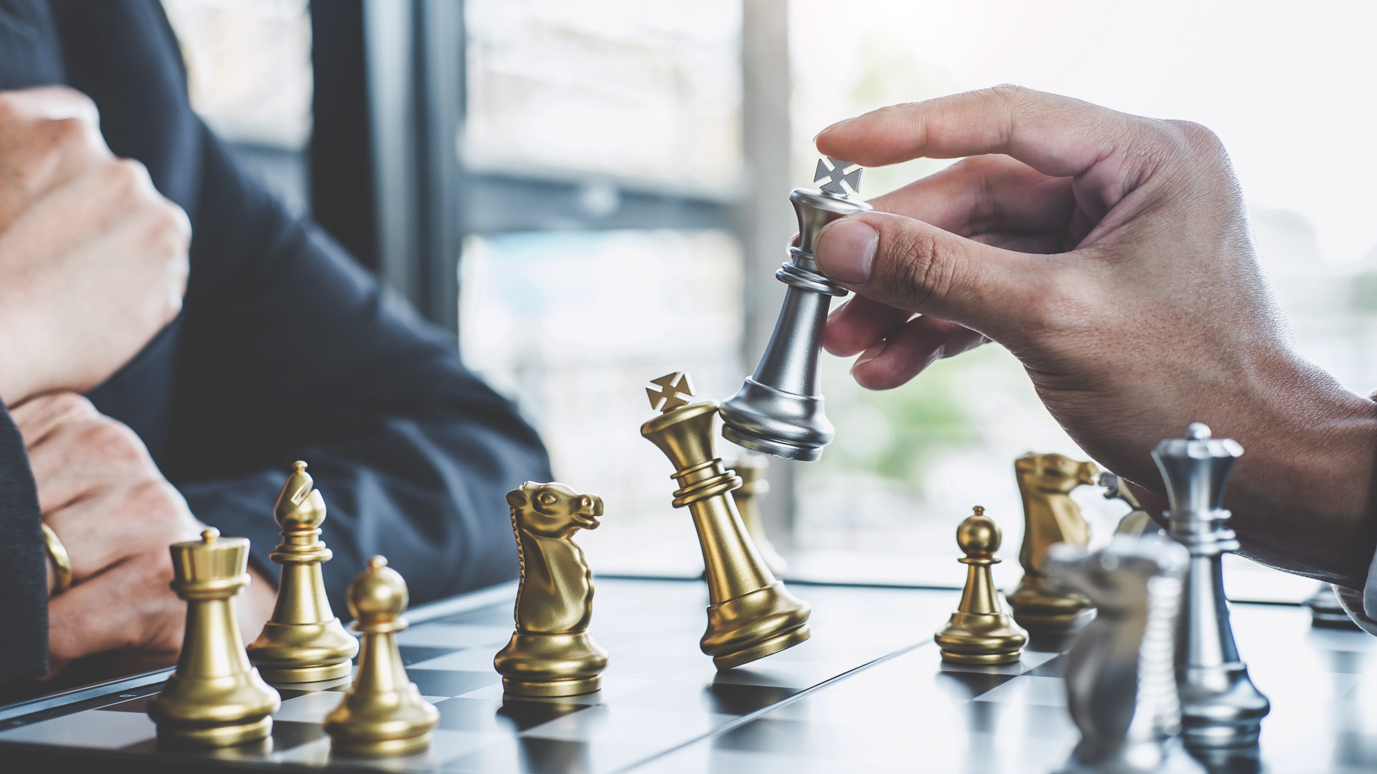 A close-up of a chess game in progress. A hand is holding a silver king piece above the board, surrounded by various gold and silver chess pieces. The setting appears to be in an office or indoors with natural light coming through a window.