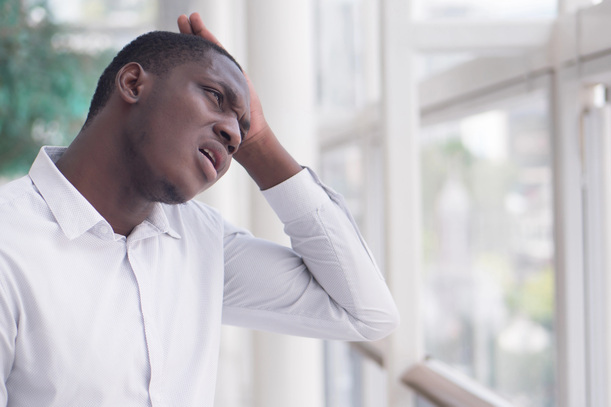 A man in a white shirt looks upset or stressed, holding his head with one hand. He is standing indoors near a window with a blurred view of greenery outside.