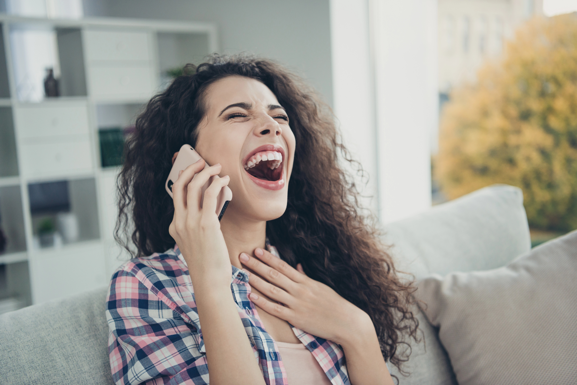 A woman with curly hair, wearing a plaid shirt, is sitting on a couch, holding a smartphone to her ear. She appears to be laughing or excited, with one hand on her chest. The background shows a bright interior with a window and shelves.