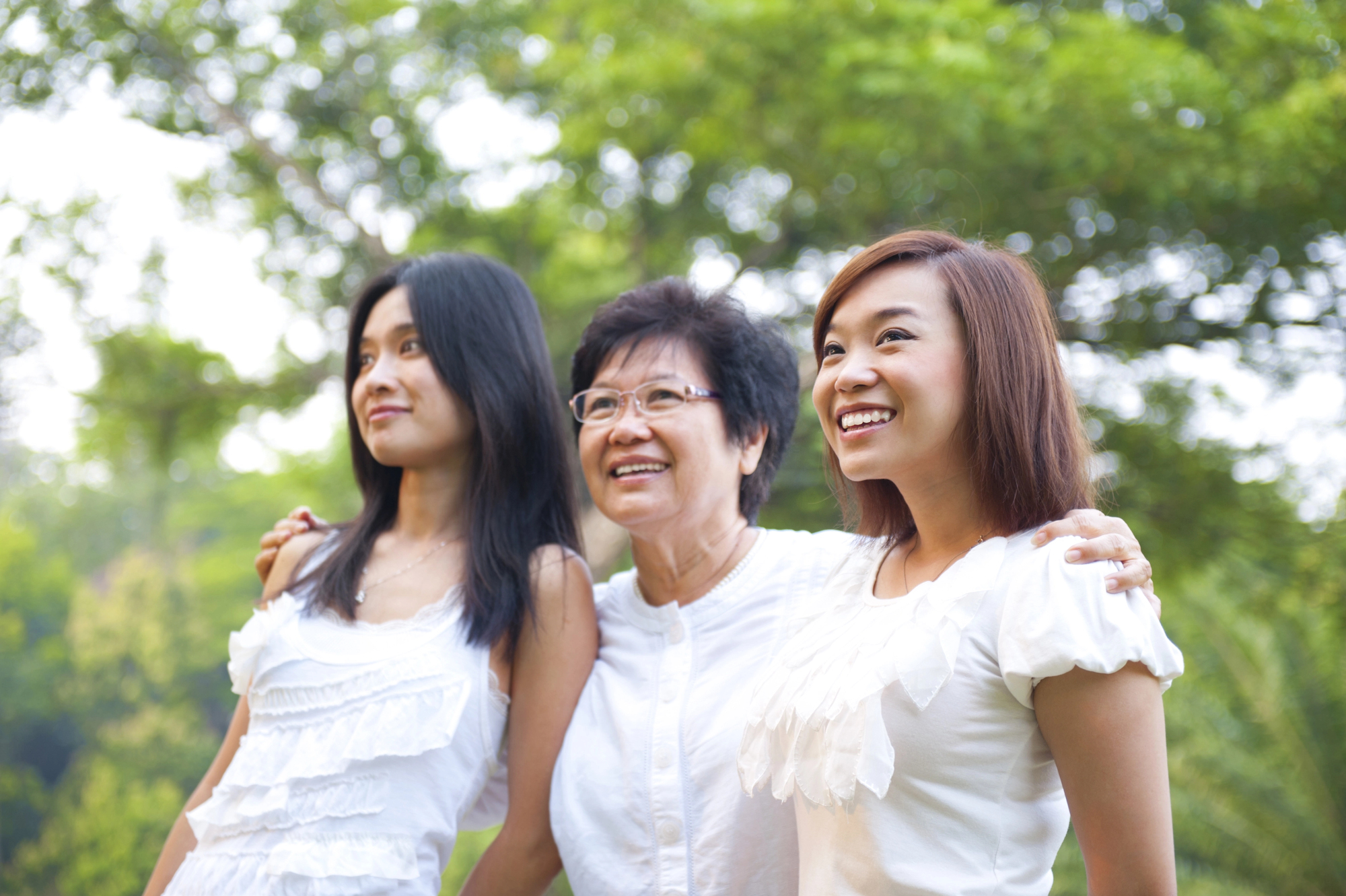 Three women stand closely together in a park, smiling and looking ahead. They are wearing white tops, and the background is lush with green trees, suggesting a bright, sunny day.