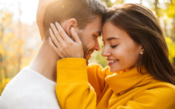 A couple embraces closely in a sunlit outdoor setting, both smiling with eyes closed. The woman wears a bright yellow jacket, while the man wears a white shirt. Sunlight filters through trees in the background, creating a warm, serene atmosphere.