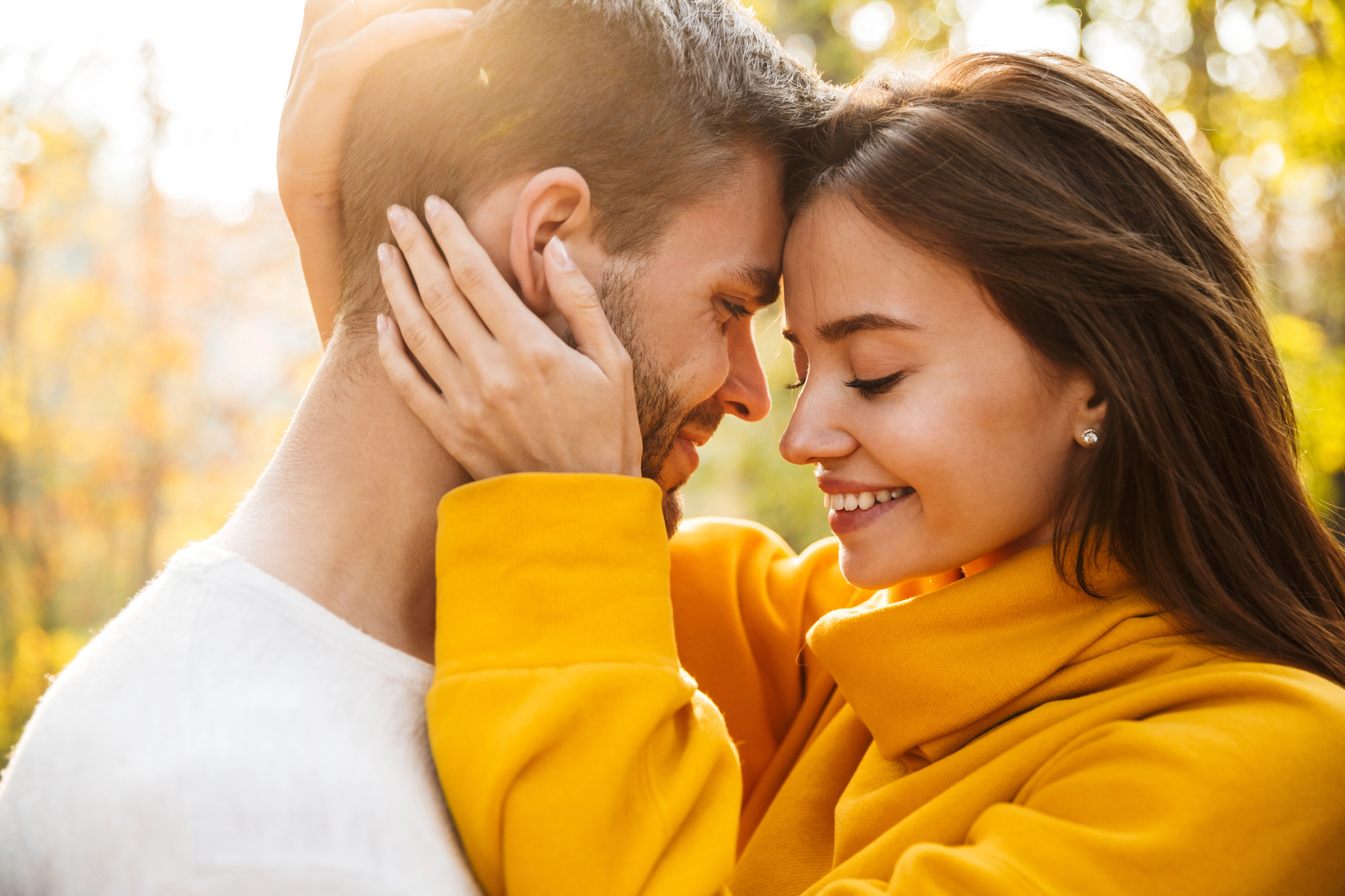 A couple embraces closely in a sunlit outdoor setting, both smiling with eyes closed. The woman wears a bright yellow jacket, while the man wears a white shirt. Sunlight filters through trees in the background, creating a warm, serene atmosphere.