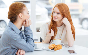 Two women sit at a table in a cafe, engaged in conversation. Both have cups of coffee in front of them, and two croissants are on a plate. One woman gestures while speaking. A smartphone lies on the table. Bright windows in the background.