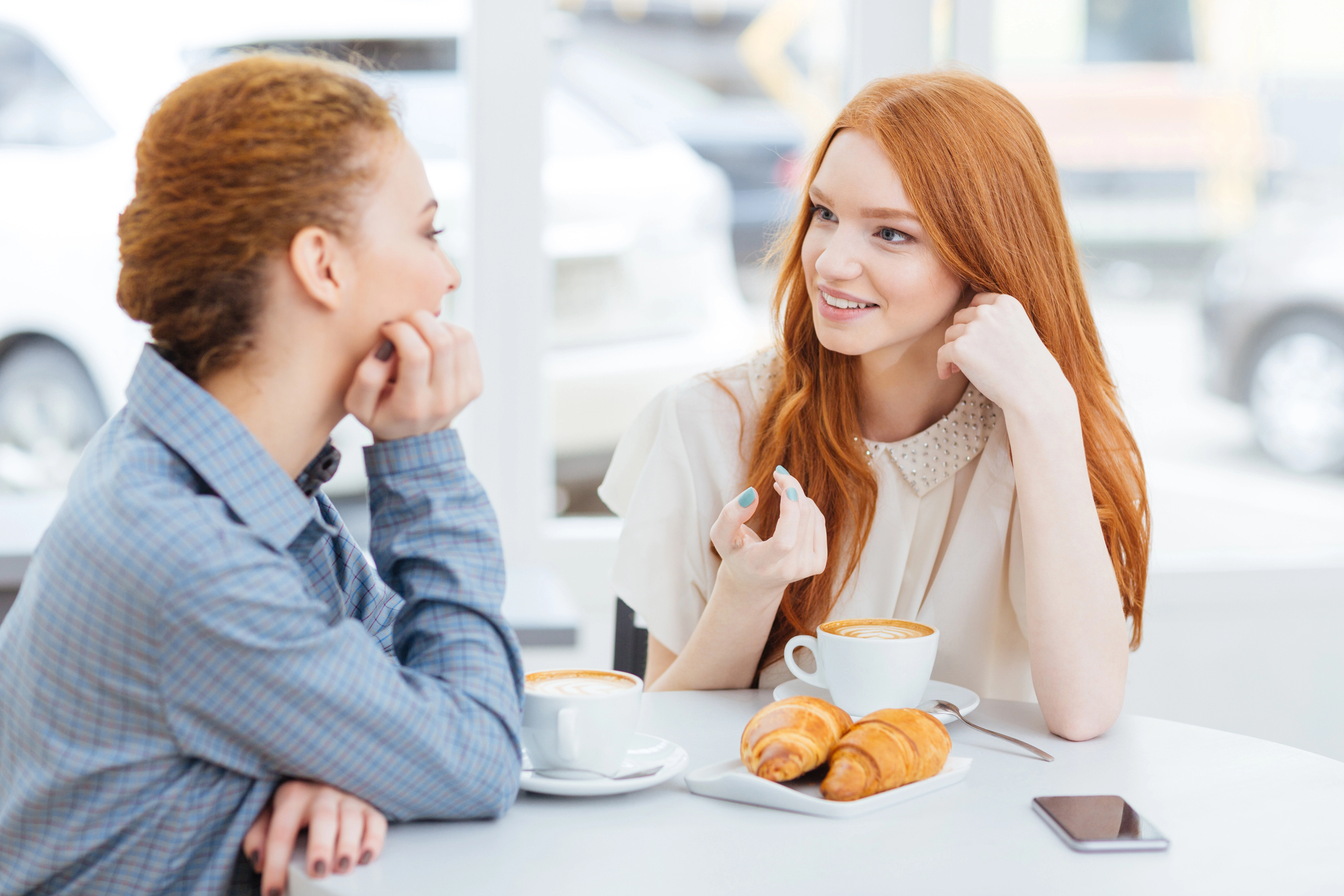 Two women sit at a table in a cafe, engaged in conversation. Both have cups of coffee in front of them, and two croissants are on a plate. One woman gestures while speaking. A smartphone lies on the table. Bright windows in the background.