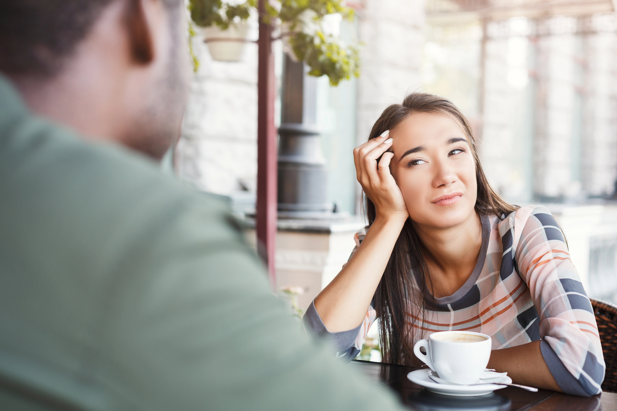 A woman sitting at an outdoor café, resting her head on her hand and looking thoughtfully at a man across from her. A cup of coffee is on the table in front of her. The background shows a blurred street scene with plants.