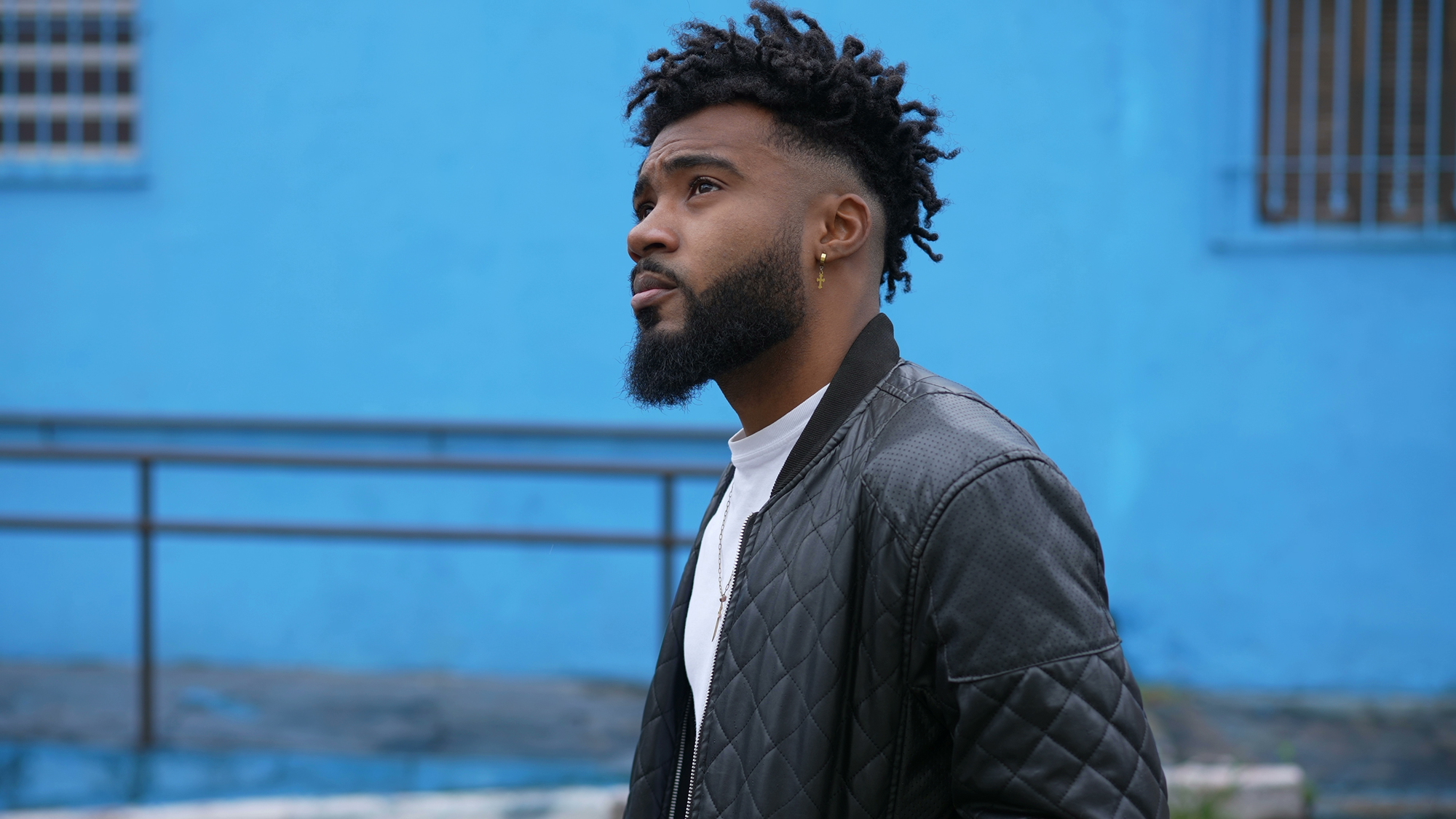 A person with curly hair and a beard stands outdoors, looking off into the distance. They are wearing a black quilted jacket over a white shirt. The background features a vibrant blue wall and blurred railing.