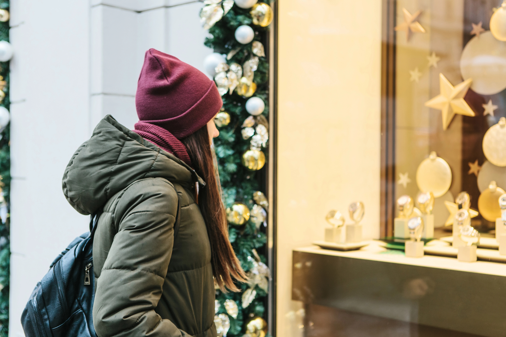 A person in a green coat and maroon beanie looks at a decorated shop window display featuring gold and white ornaments. The background includes festive garlands with baubles.