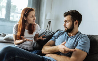 A woman and a man are sitting on a couch in a living room, engaged in a heated discussion. The woman gestures with her hands, looking frustrated, while the man, with crossed arms and an incredulous expression, responds defensively.