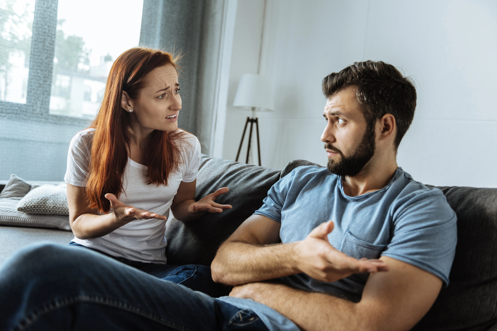 A woman and a man are sitting on a couch in a living room, engaged in a heated discussion. The woman gestures with her hands, looking frustrated, while the man, with crossed arms and an incredulous expression, responds defensively.