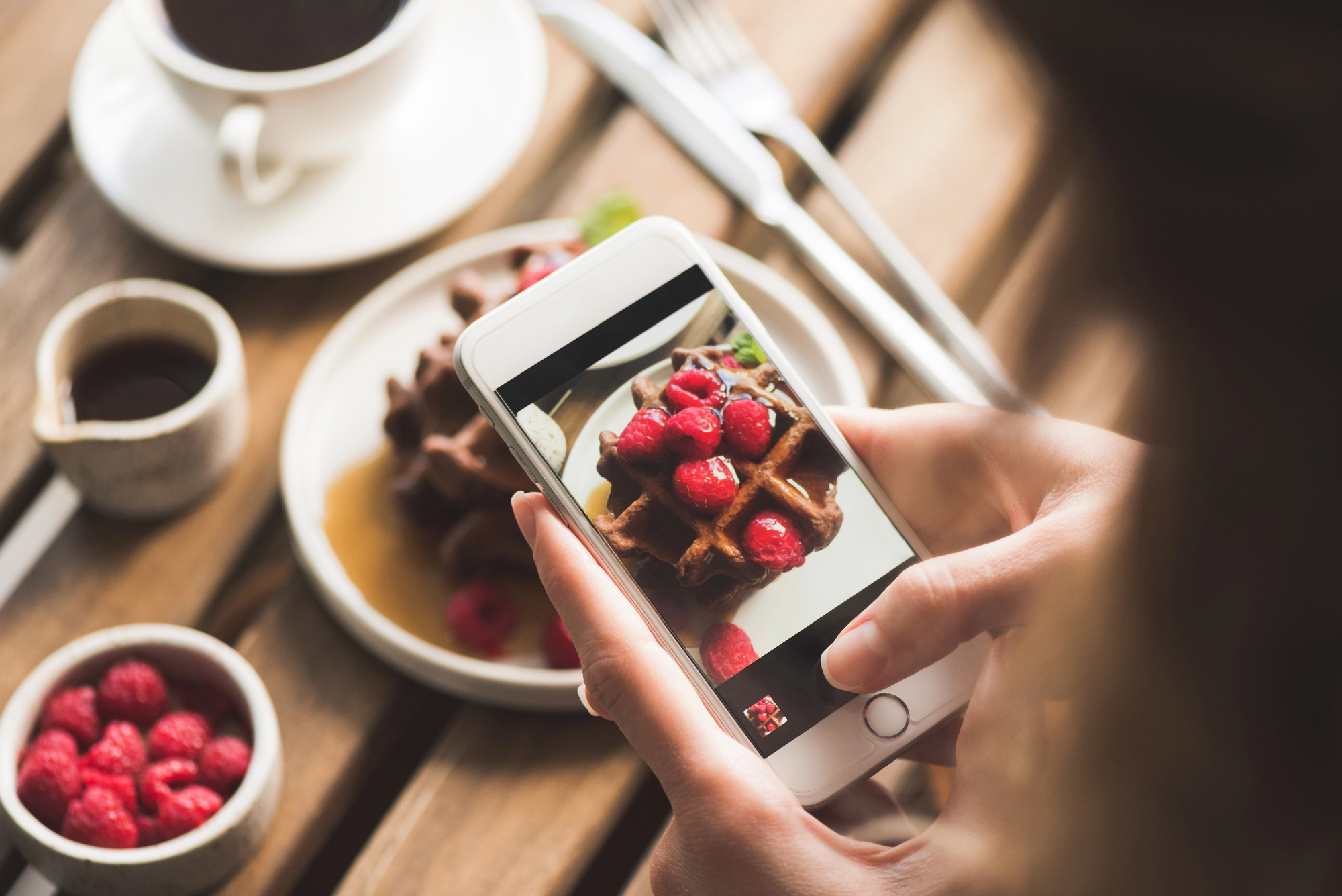 A person taking a photo of a plate with waffles topped with raspberries using a smartphone. There's a cup of coffee and a small jug on the wooden table nearby, along with extra raspberries in a bowl.