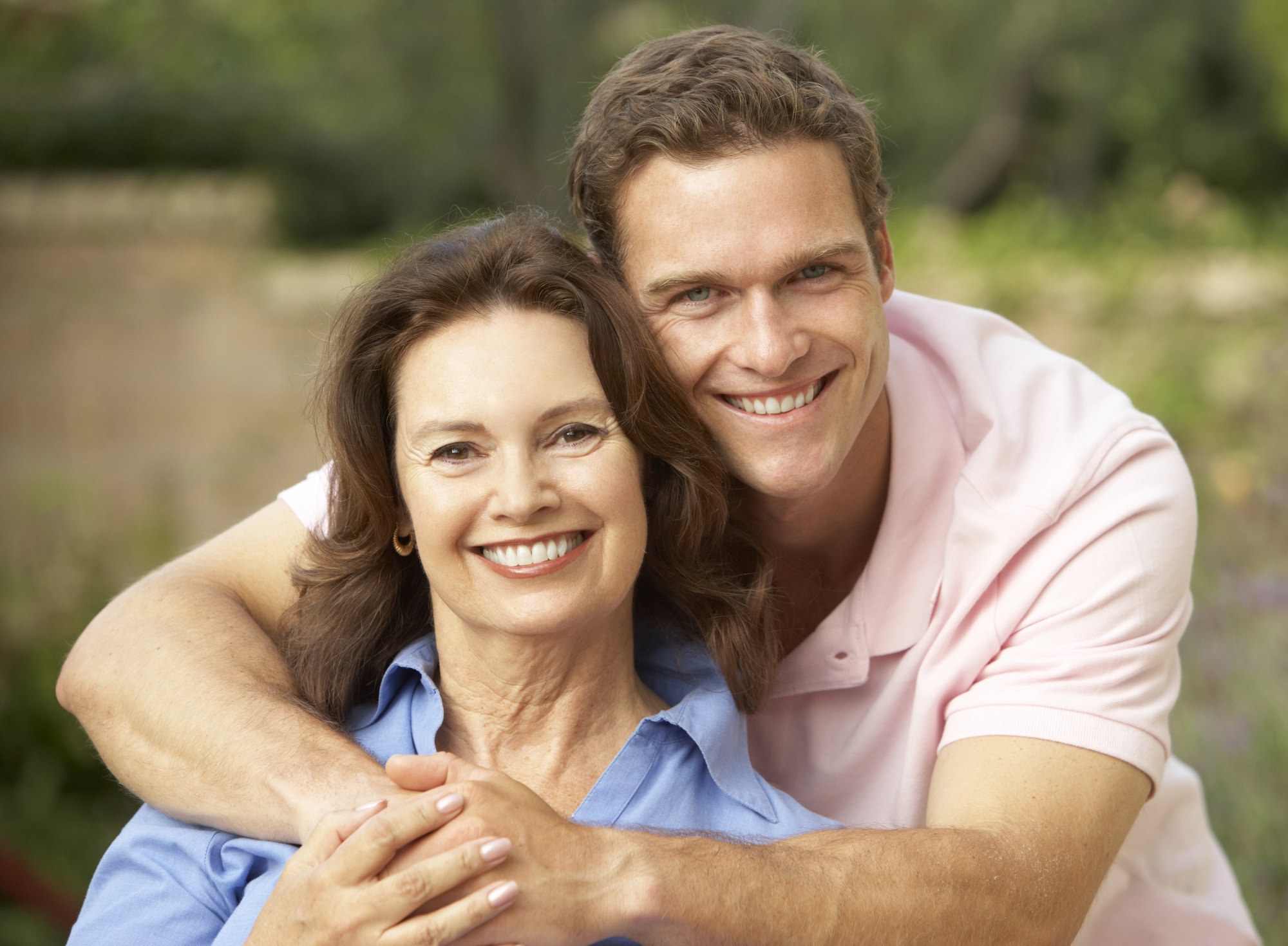 A man in a pink shirt hugs a smiling woman in a blue shirt from behind. Both are outdoors with a blurred green background. They appear cheerful and relaxed.