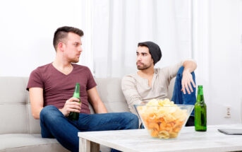 Two men sit on a sofa with beers, engaged in conversation. A bowl of chips is on the coffee table in front of them. One wears a maroon shirt and jeans; the other, a gray shirt, jeans, and a beanie. A phone rests on the table beside them.