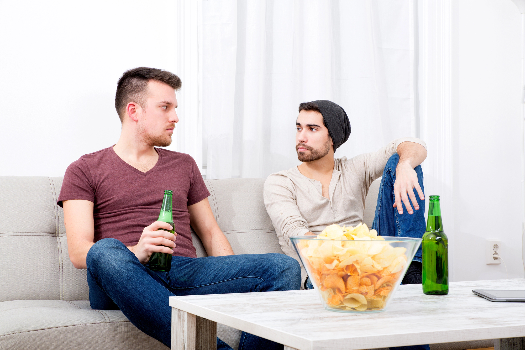 Two men sit on a sofa with beers, engaged in conversation. A bowl of chips is on the coffee table in front of them. One wears a maroon shirt and jeans; the other, a gray shirt, jeans, and a beanie. A phone rests on the table beside them.