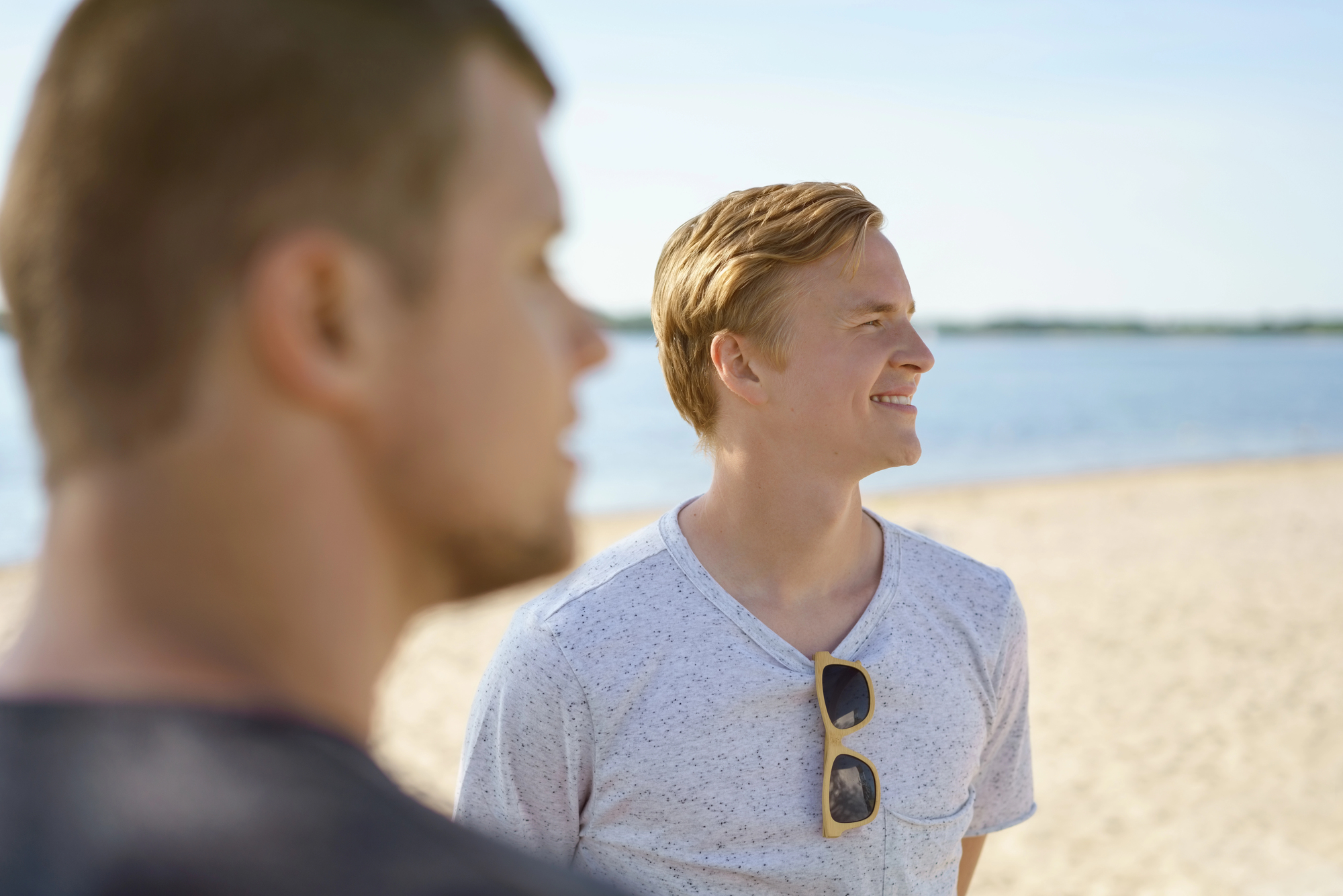 Two people at a sunny beach. One man in focus wearing a gray t-shirt and sunglasses around his neck smiles while looking into the distance. Another man in the foreground is blurred. The ocean and sky are visible in the background.