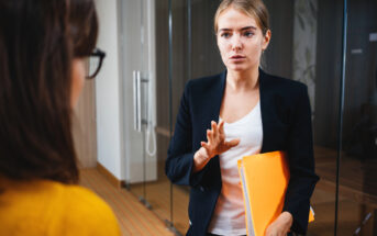 A woman in a black blazer and white shirt holds yellow folders while speaking to another person. They are standing in an office setting with glass walls. The woman appears focused and is gesturing with her hand.