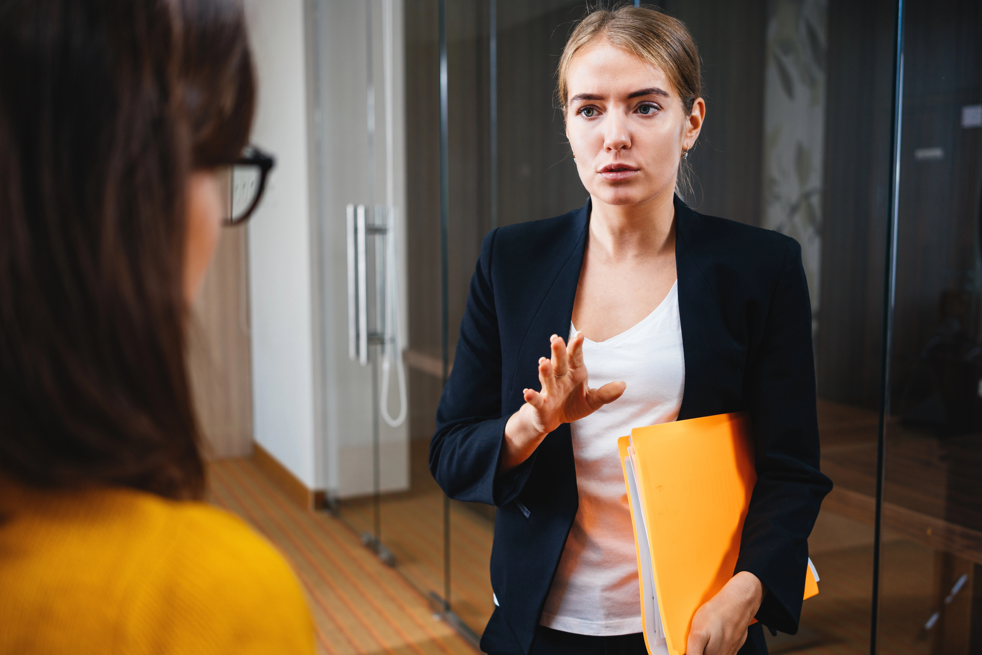 A woman in a black blazer and white shirt holds yellow folders while speaking to another person. They are standing in an office setting with glass walls. The woman appears focused and is gesturing with her hand.