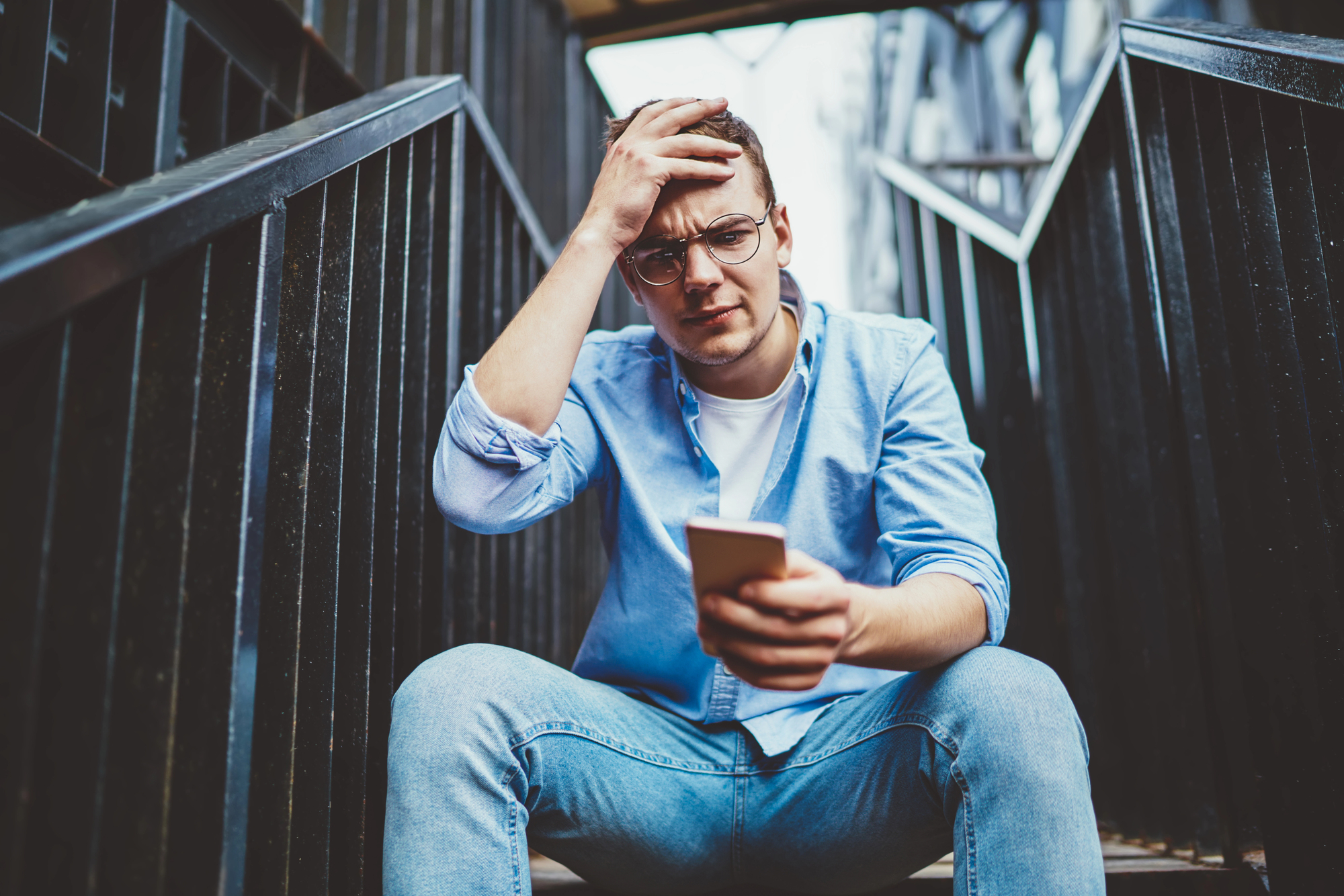 A young man with glasses, wearing a blue shirt and jeans, sits on stairs, looking frustrated while holding a smartphone. His left hand rests on his head, and the background features black stair railings.