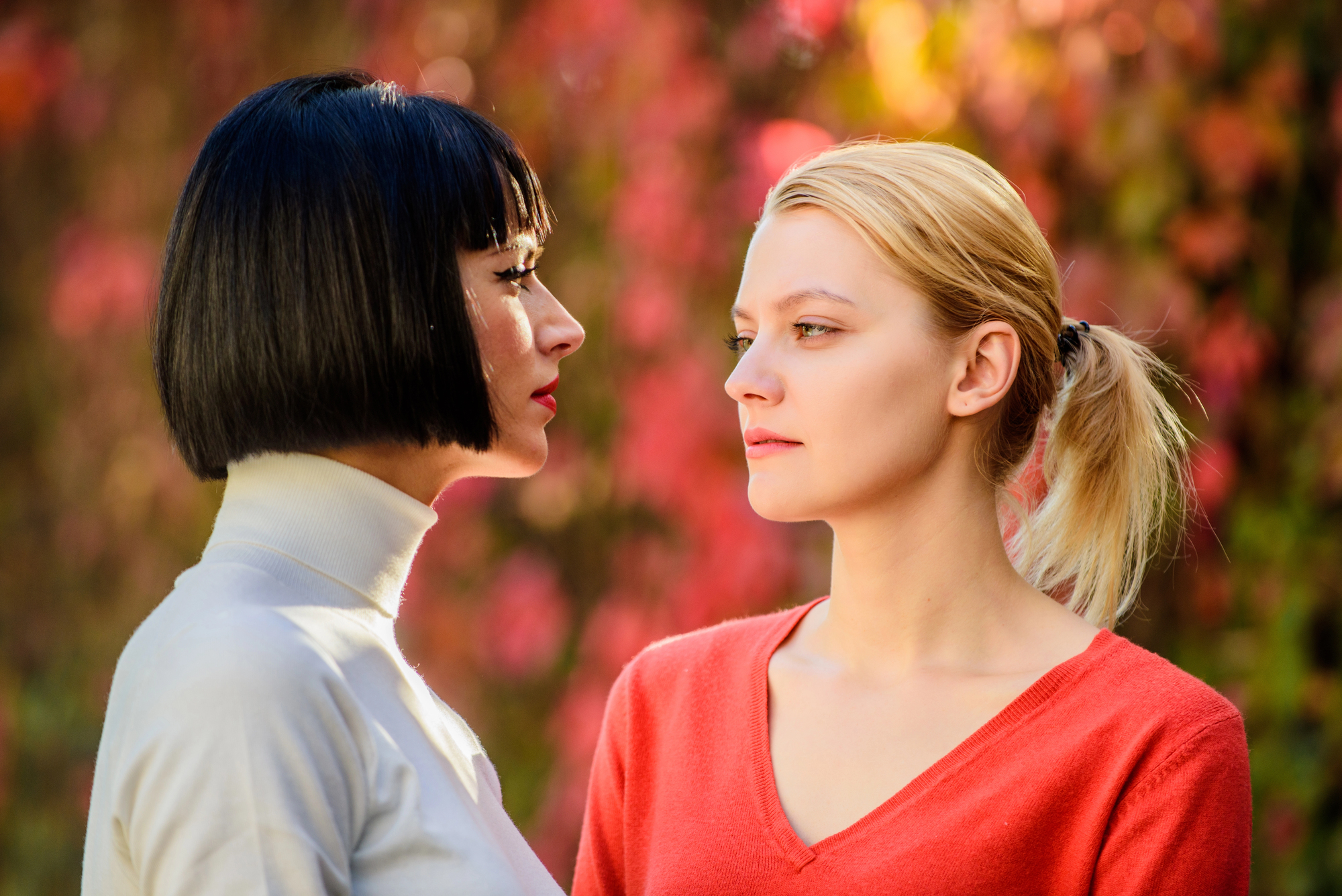 Two women face each other outdoors, with one having black hair in a bob cut and wearing a white turtleneck, and the other with blonde hair in a ponytail wearing a red sweater. The background is filled with blurred red and green foliage.