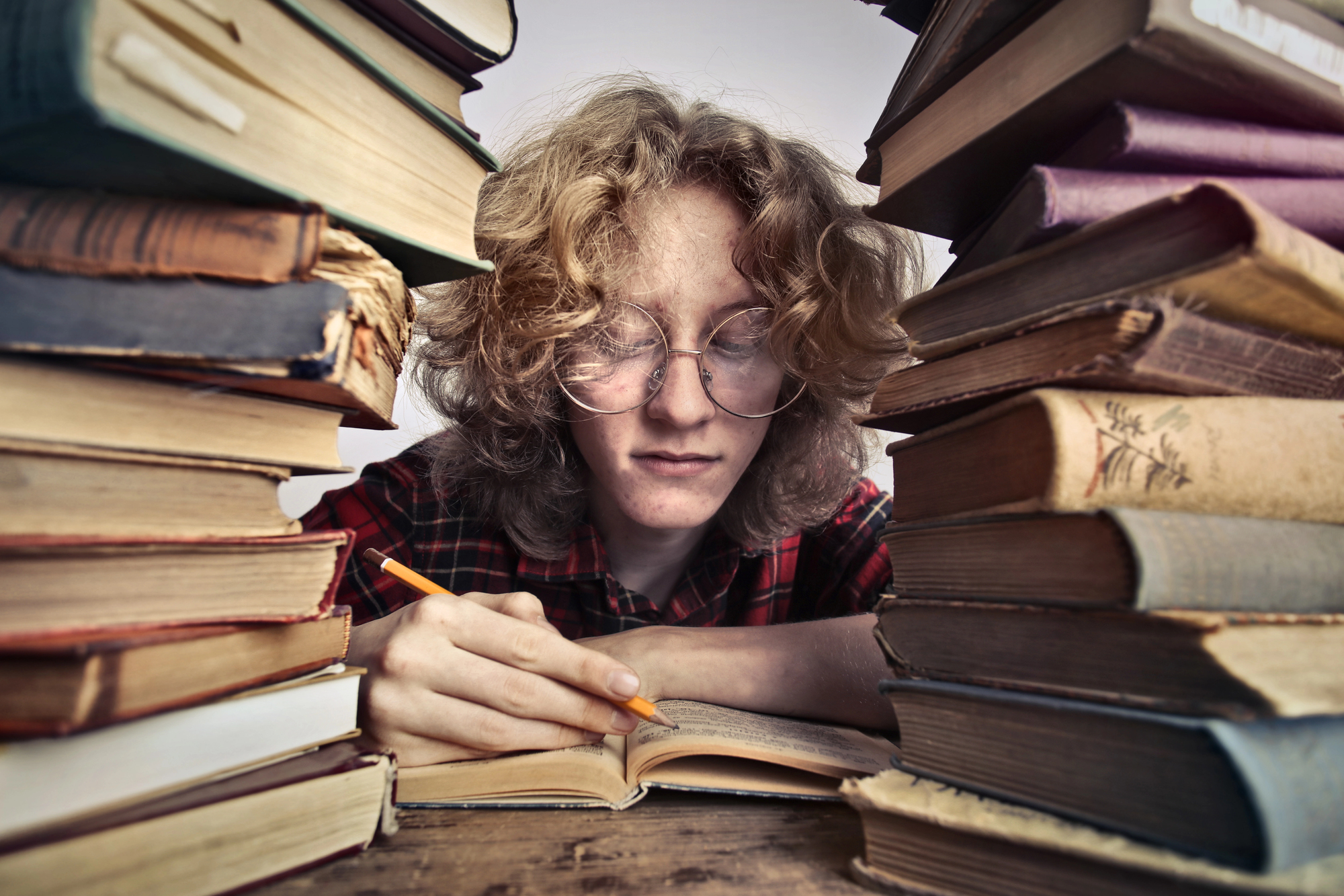 A person with curly hair and glasses is intensely focused on writing in a notebook, surrounded by stacks of books on a wooden table, suggesting a studious or academic setting.