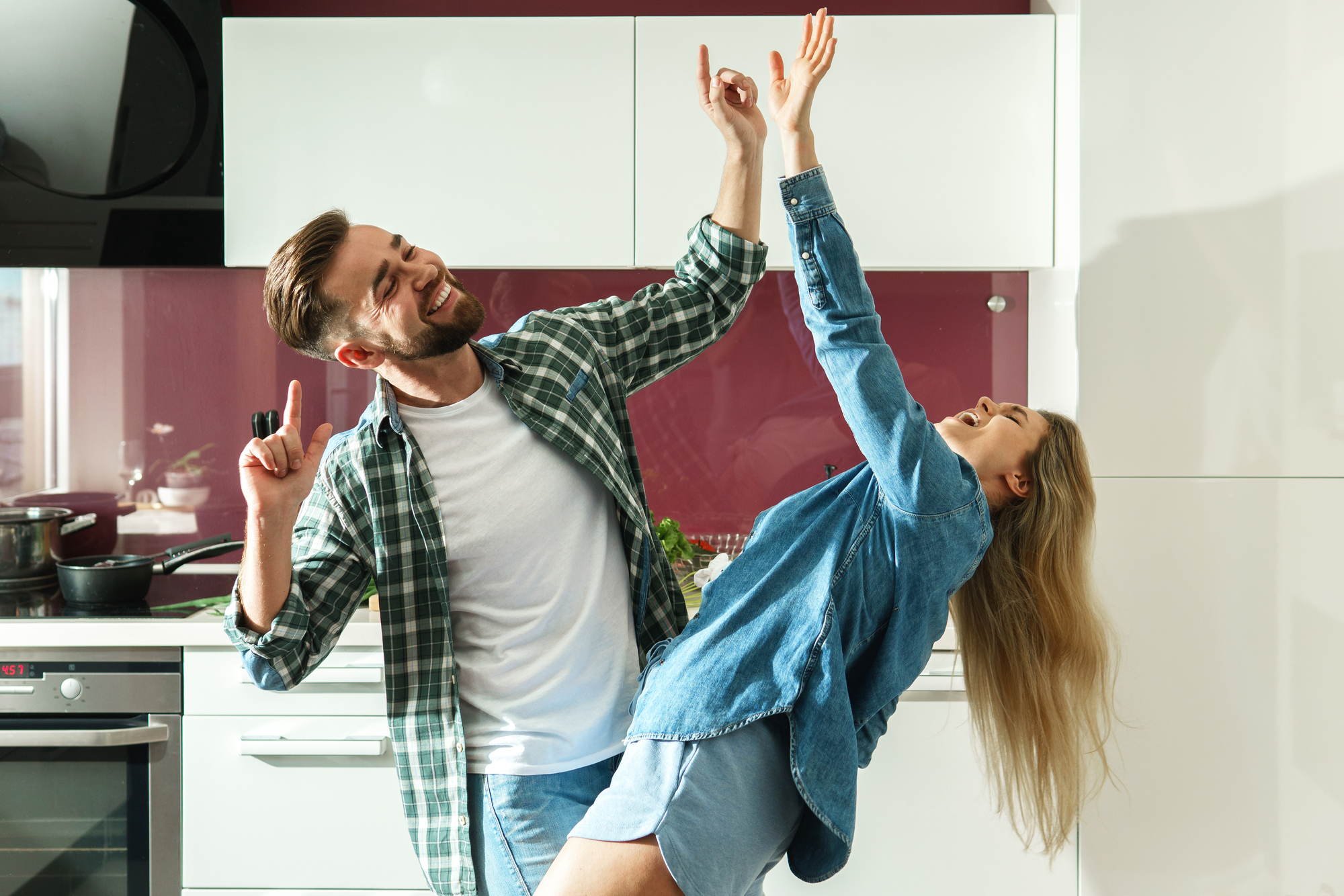 A man and woman in casual clothes dance joyfully in a modern kitchen. The woman leans back with her hand in the air, while the man points upward with a smile. The kitchen has white cabinets and a red backsplash.