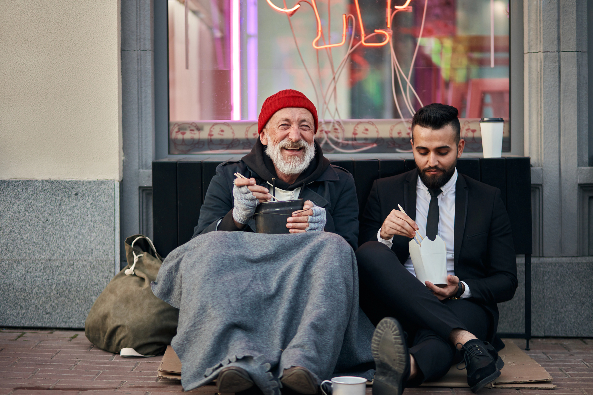 An elderly man in a red beanie, wrapped in a gray blanket, smiles while eating from a pot. A young man in a suit sits beside him, eating from a takeout box. They are sitting on a city sidewalk with a neon sign in the background.