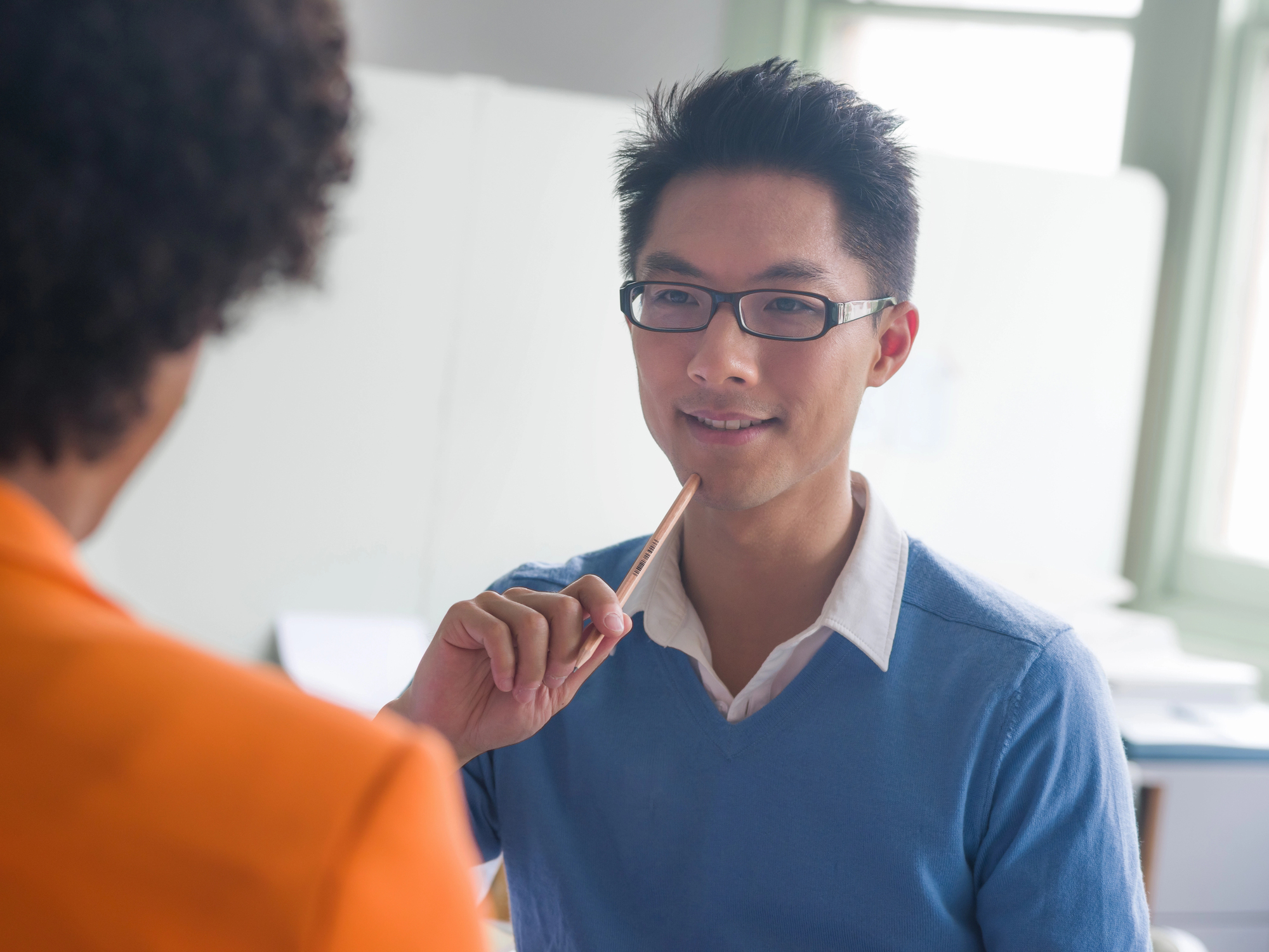 A person wearing glasses and a blue sweater is seated, holding a pencil and smiling at another person in an orange blazer, who is turned away. They appear to be engaged in conversation in a well-lit room.