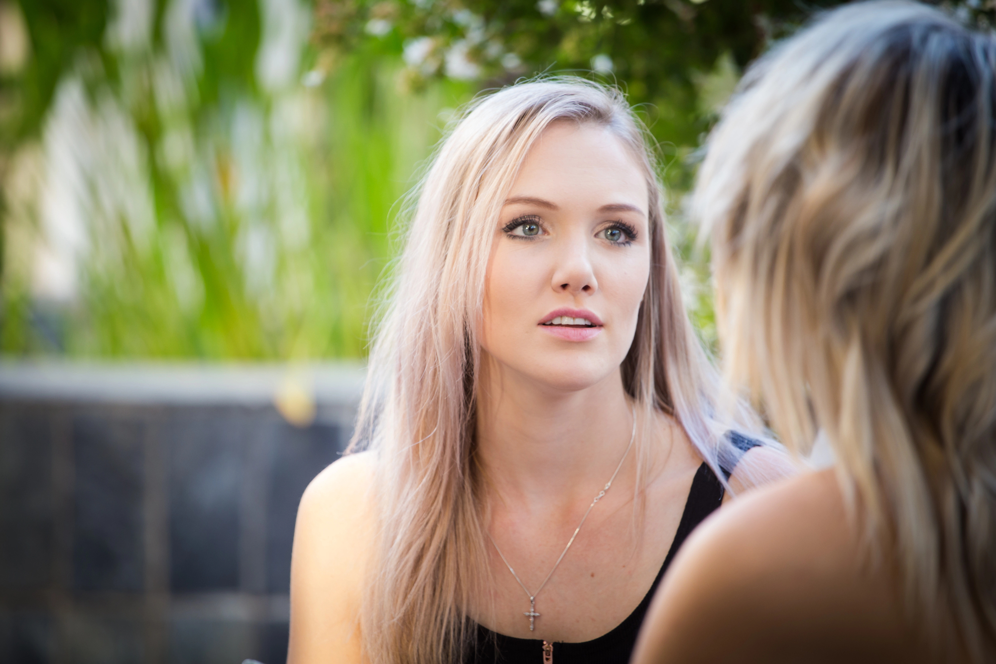 A woman with long blonde hair is sitting outdoors, engaged in conversation with another person. She is wearing a black top and a necklace with a cross pendant. The background is blurred with greenery.