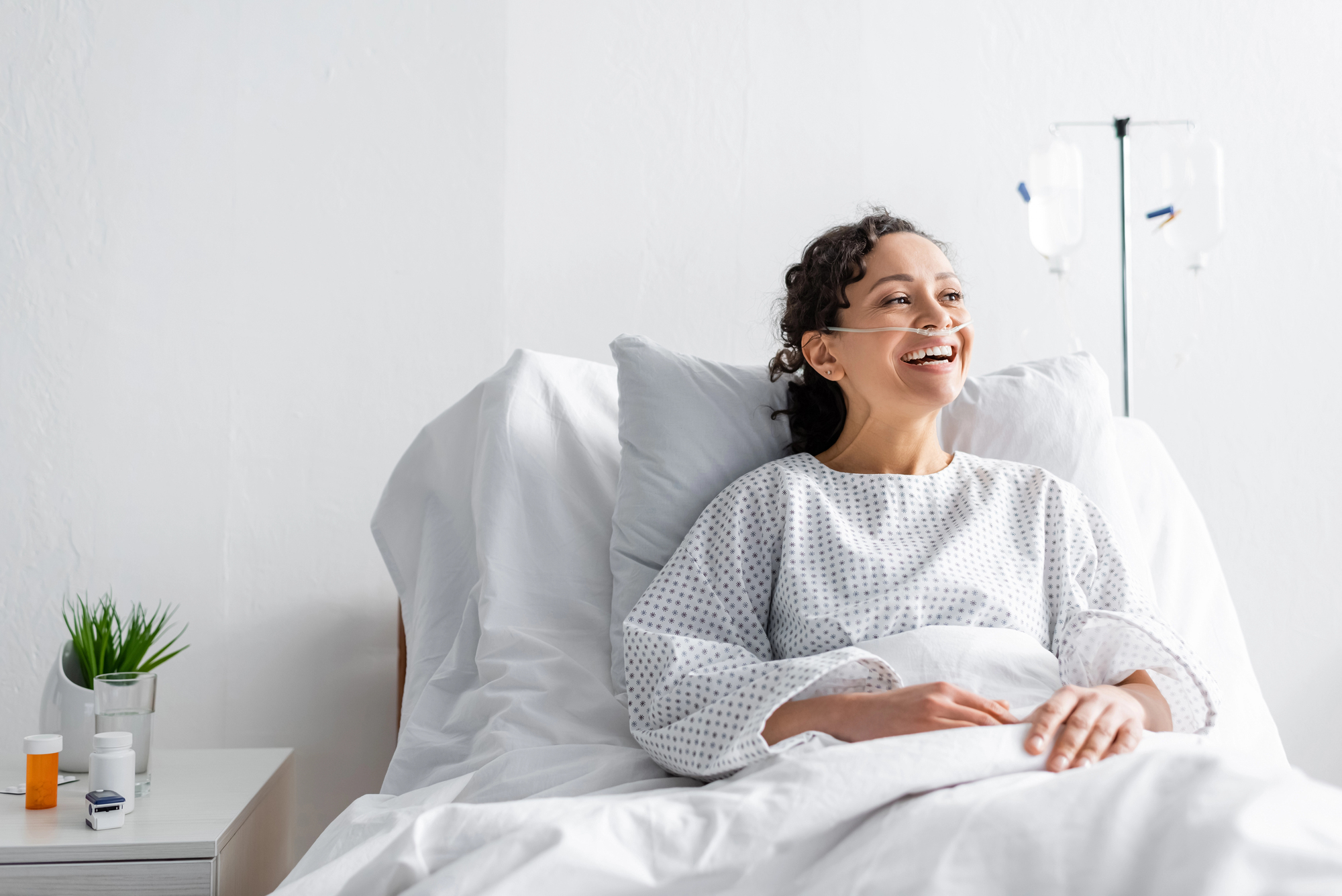 A smiling person in a hospital bed, wearing a medical gown and nasal oxygen tube. An IV stand is next to the bed. A small table with a plant and medication is beside them in a bright, clean room.