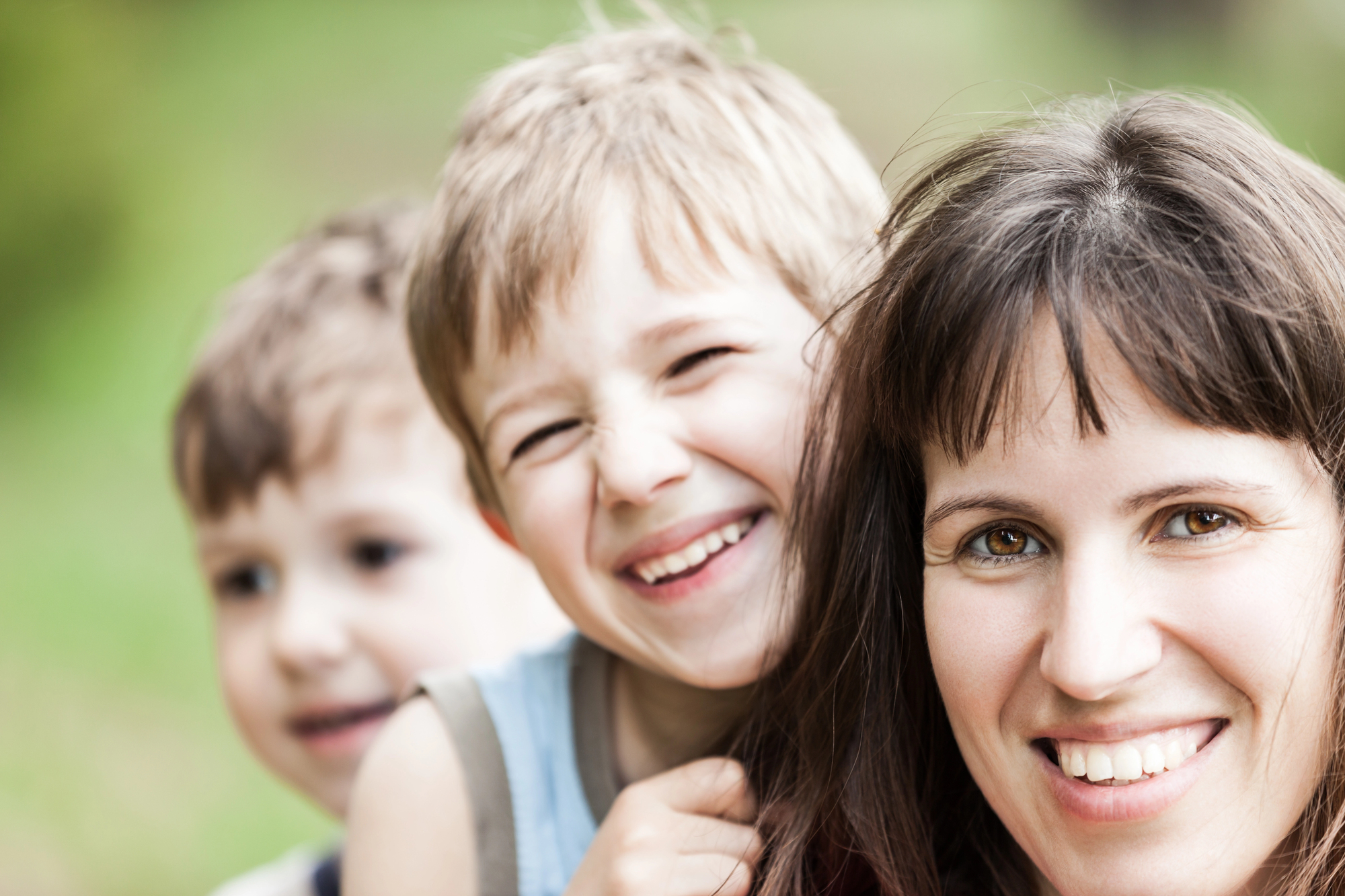 A woman and two young boys are smiling joyfully outdoors. The woman and one boy are in focus, with the background blurred, suggesting a sunny day in a park or garden. All appear happy and relaxed.