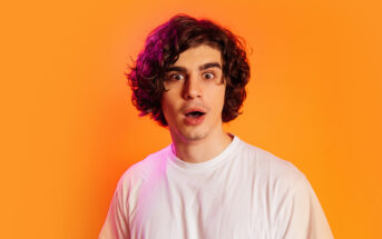 A young man with curly hair and a surprised expression stands against an orange background, wearing a white t-shirt.